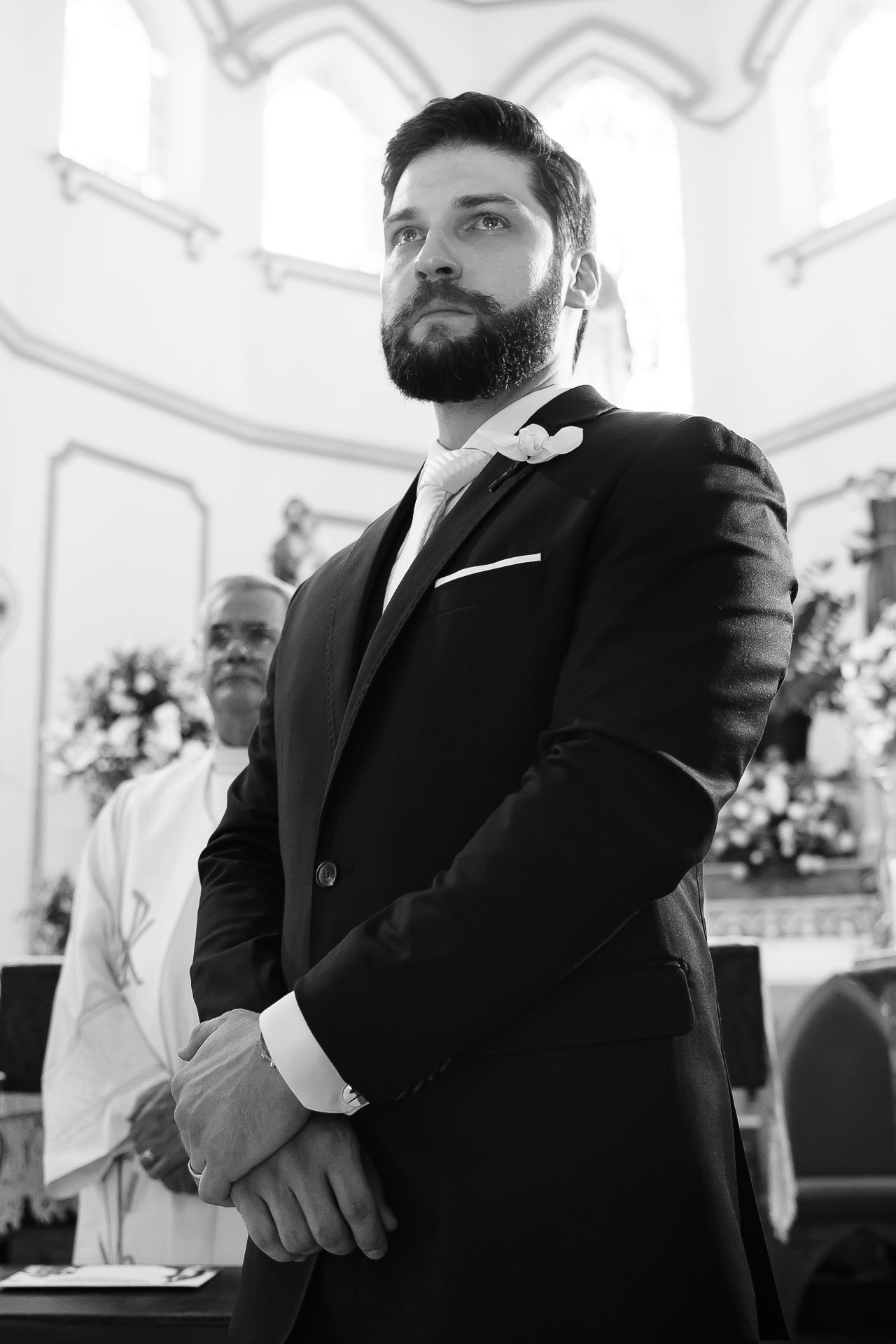 Black and white photo of a bearded groom in a suit with boutonniere standing at the altar during a church wedding ceremony.