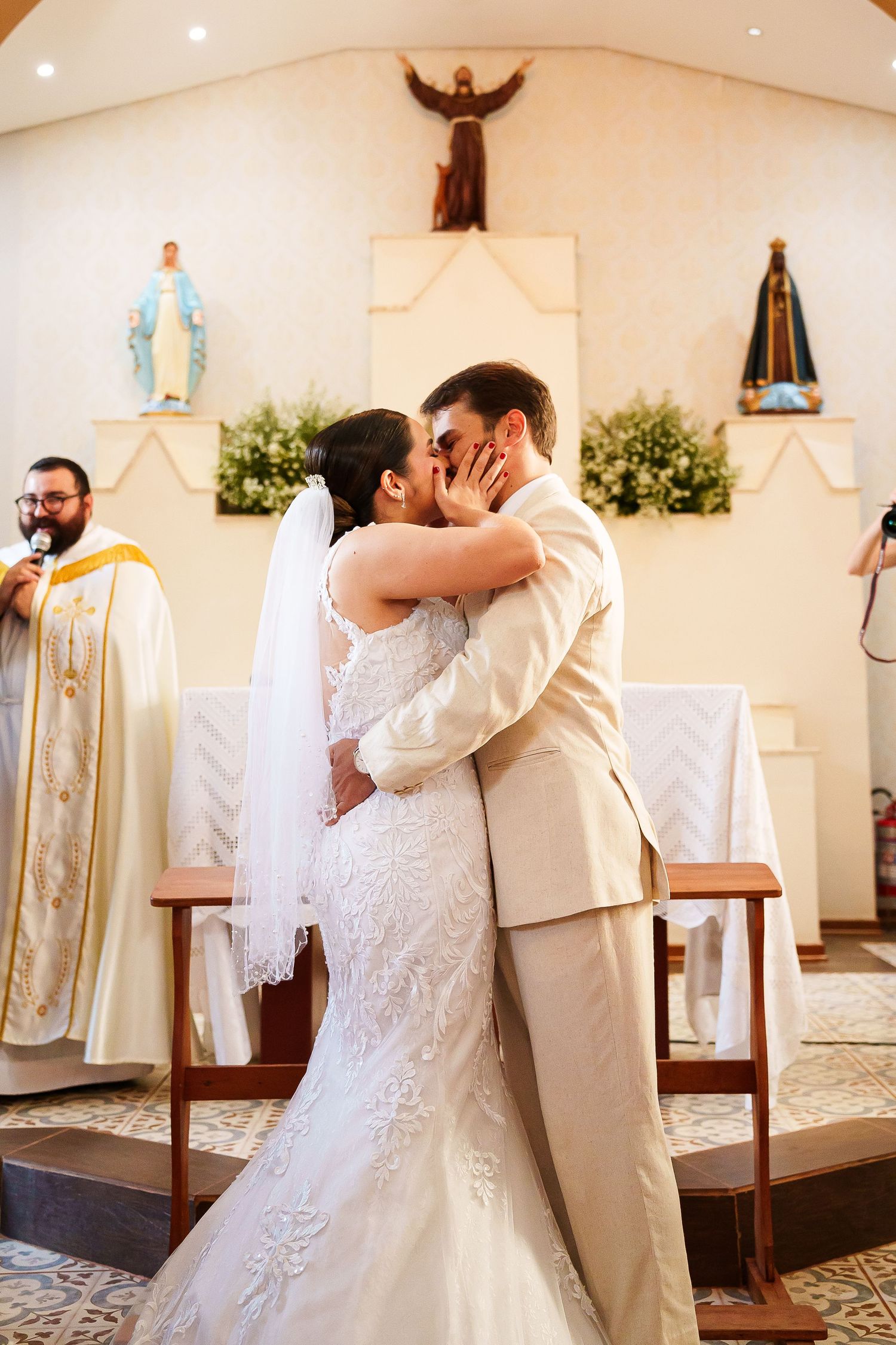 Bride and groom share first kiss at Catholic church altar during wedding ceremony, priest officiating in background.