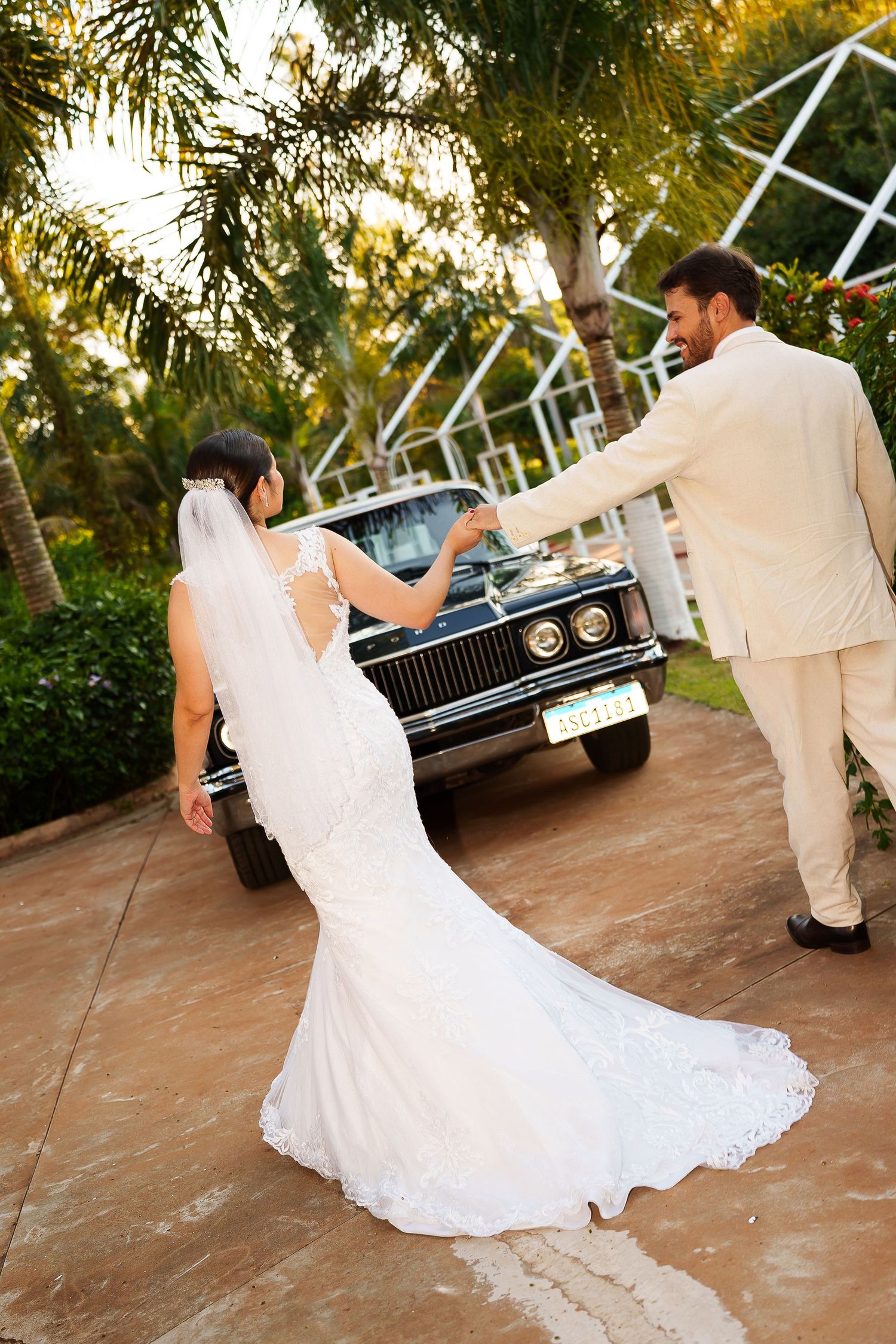 Bride and groom in white attire walk hand-in-hand toward a vintage black car surrounded by tropical palm trees.