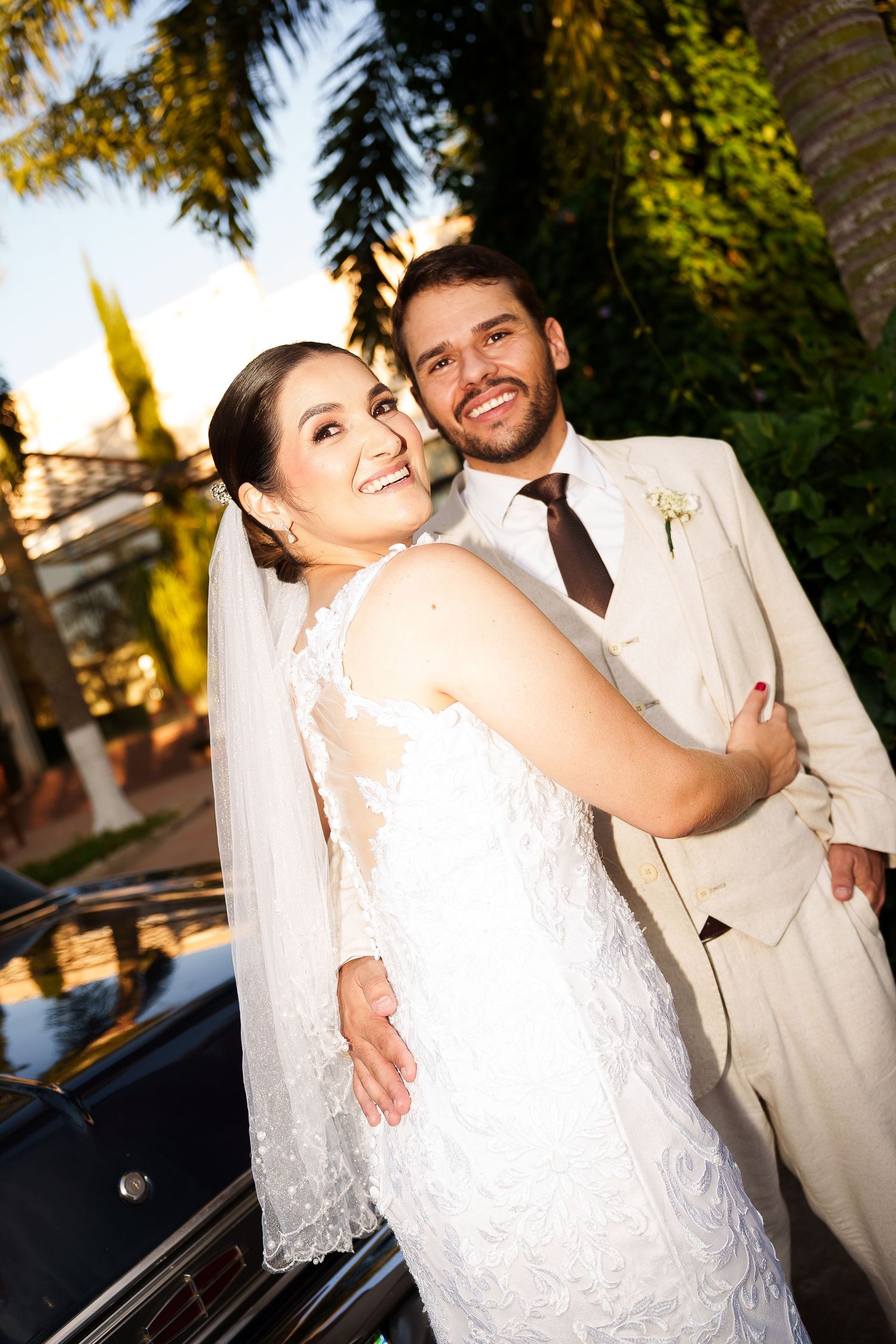 A smiling bride in a lace wedding dress and veil embraces a groom in a beige suit outdoors near palm trees.
