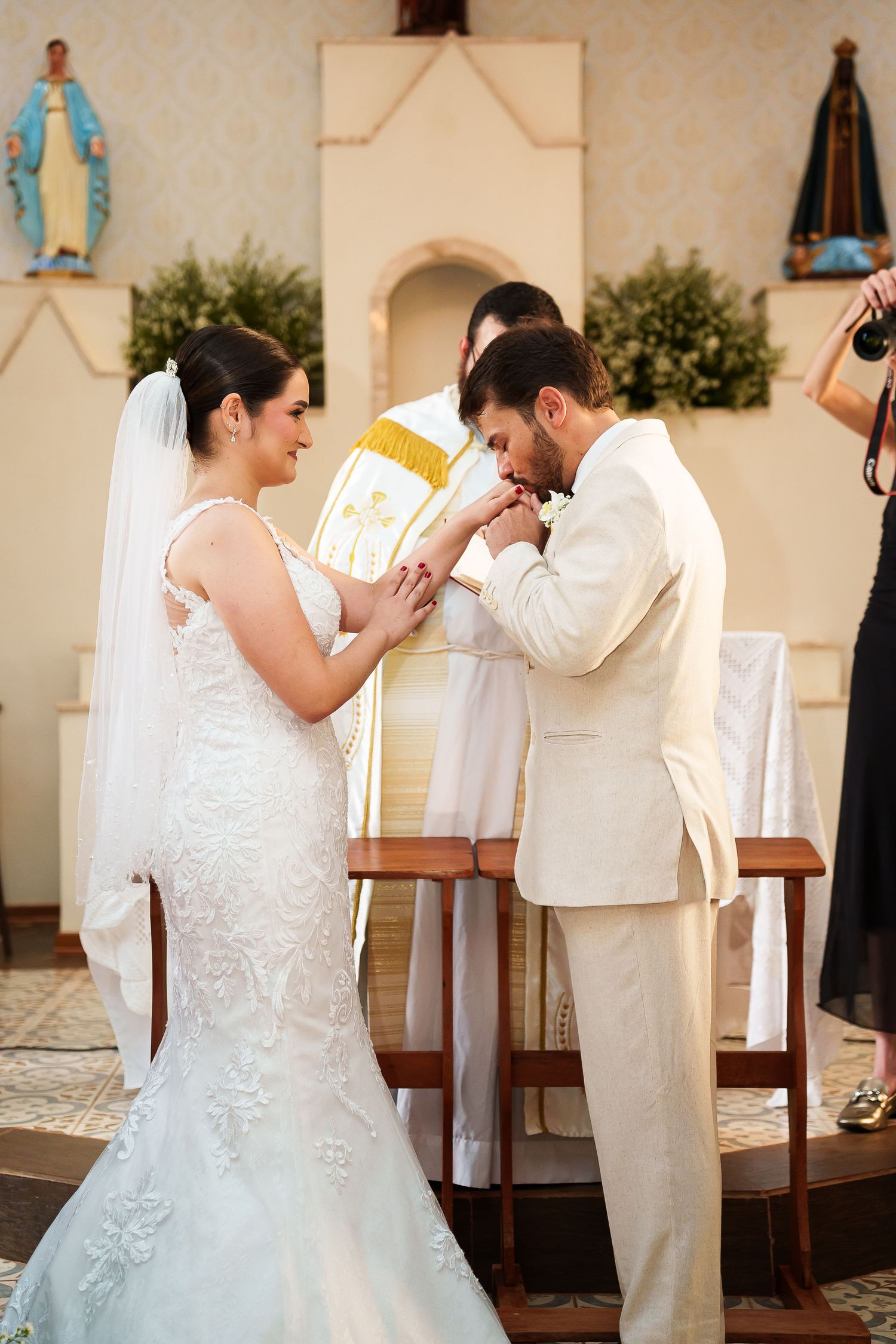 Groom kissing bride's hand at church altar during wedding ceremony, with priest in gold vestments behind them.