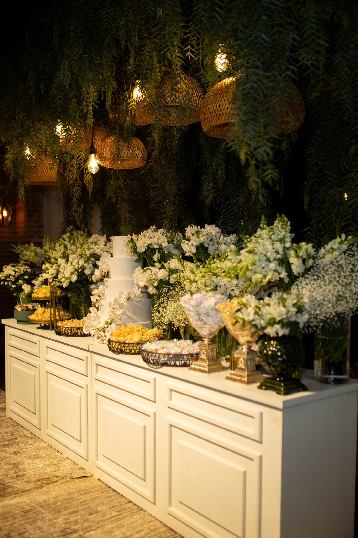 Elegant wedding dessert table with white flowers, tiered cake, and warm basket pendant lights against lush greenery.