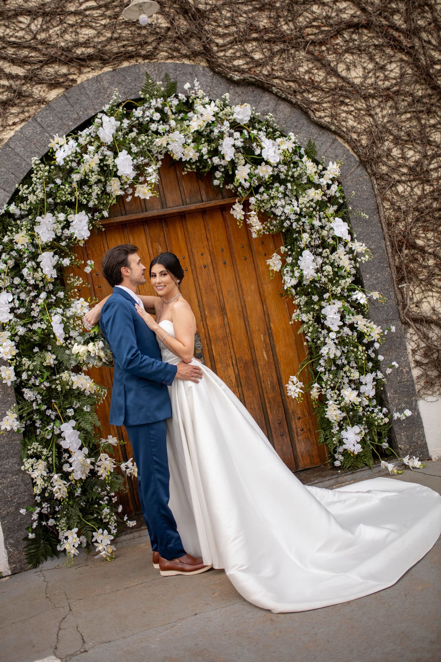 Bride and groom embracing under a white floral arch at a wooden door on their wedding day.