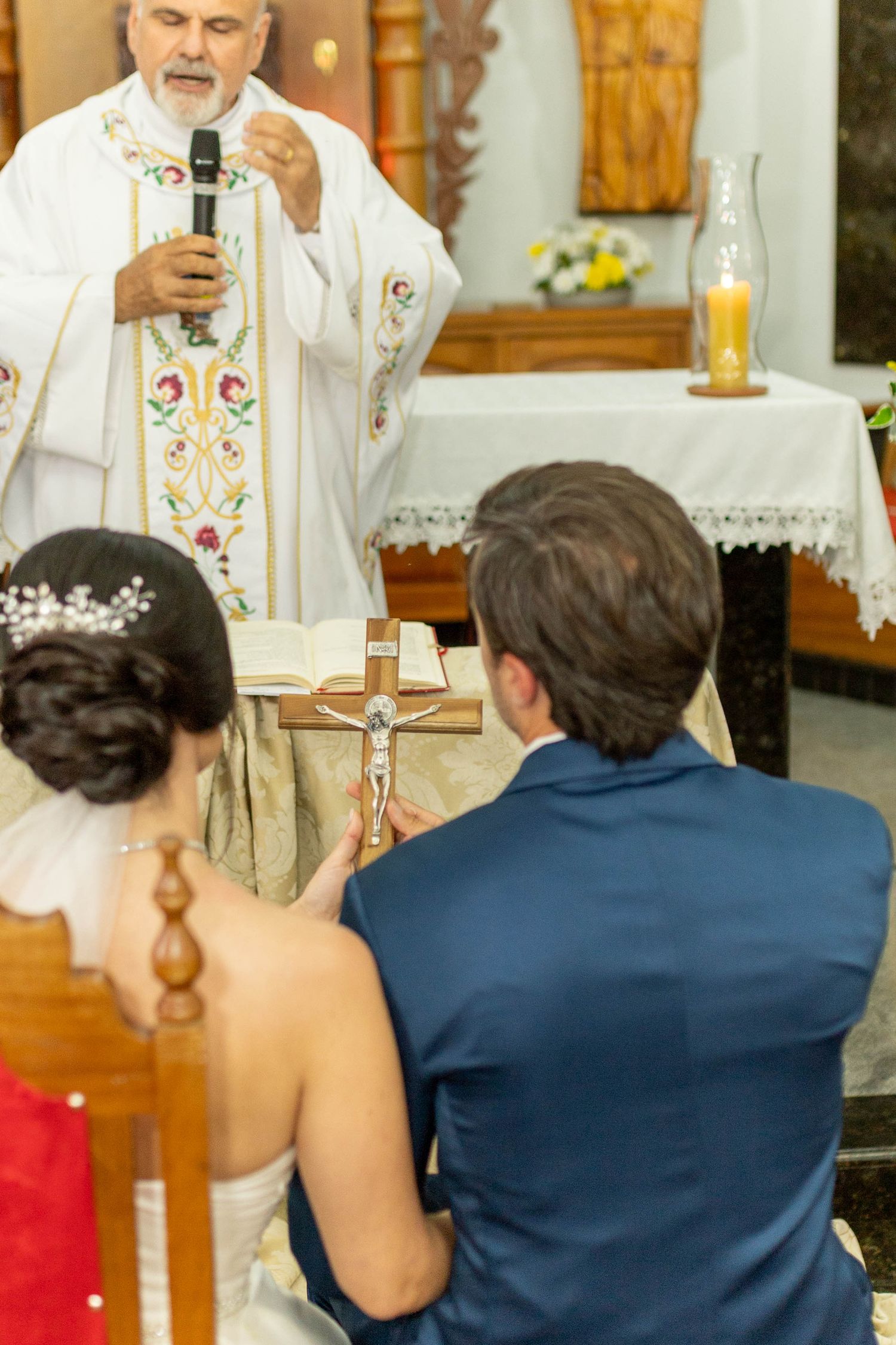 Catholic priest officiates wedding ceremony as bride and groom kneel before altar with crucifix in church.