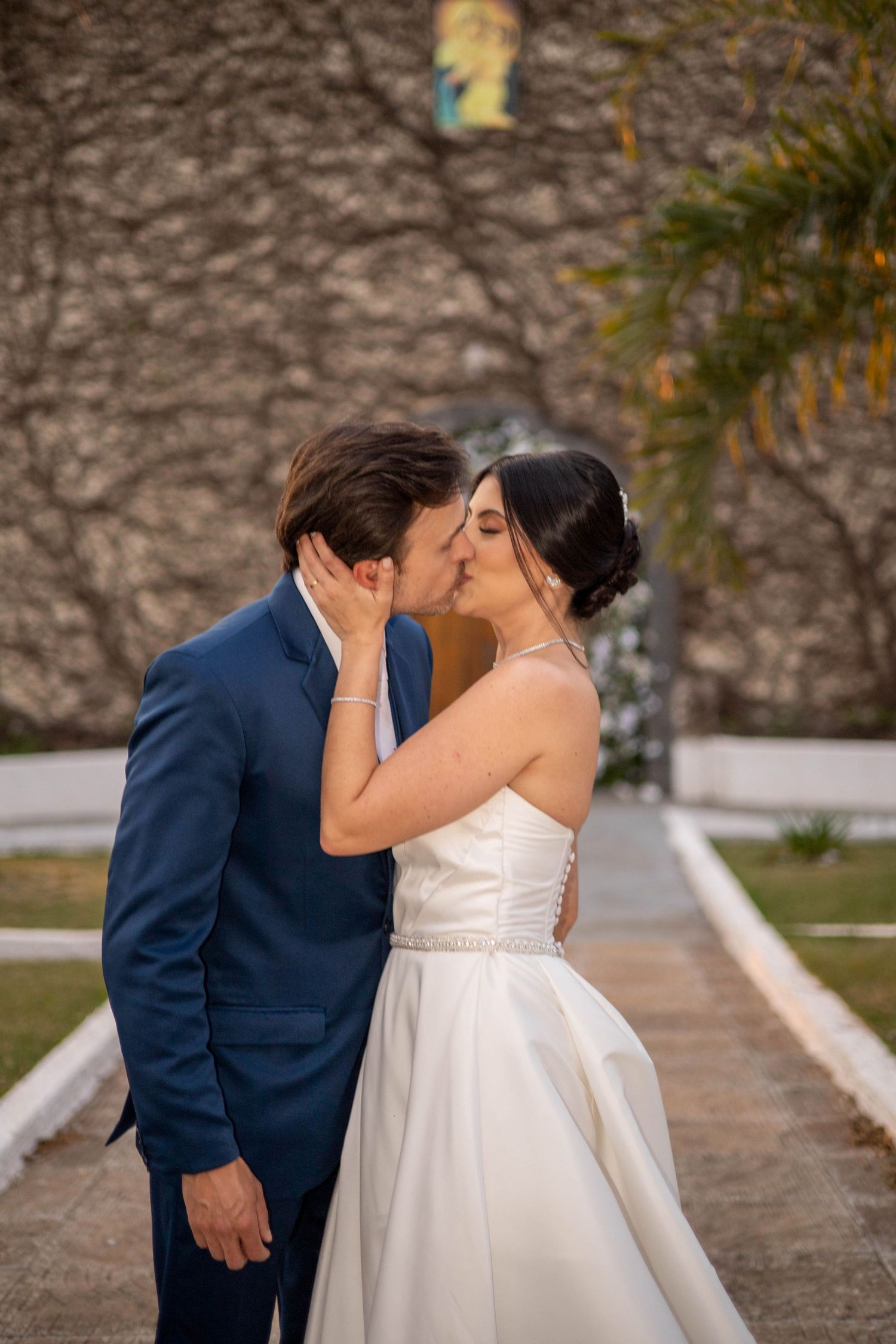 A bride and groom share a romantic kiss outdoors, she in a white gown and he in a navy suit, with a stone wall behind.