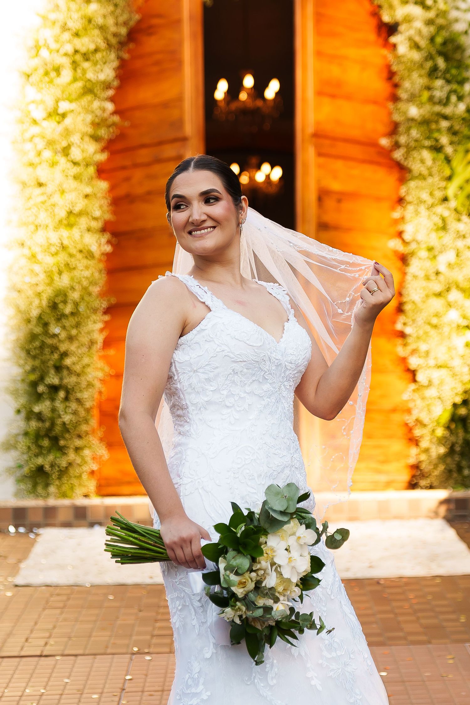 Smiling bride in white lace gown holding bouquet, lifting veil outdoors near wooden door with floral decor.