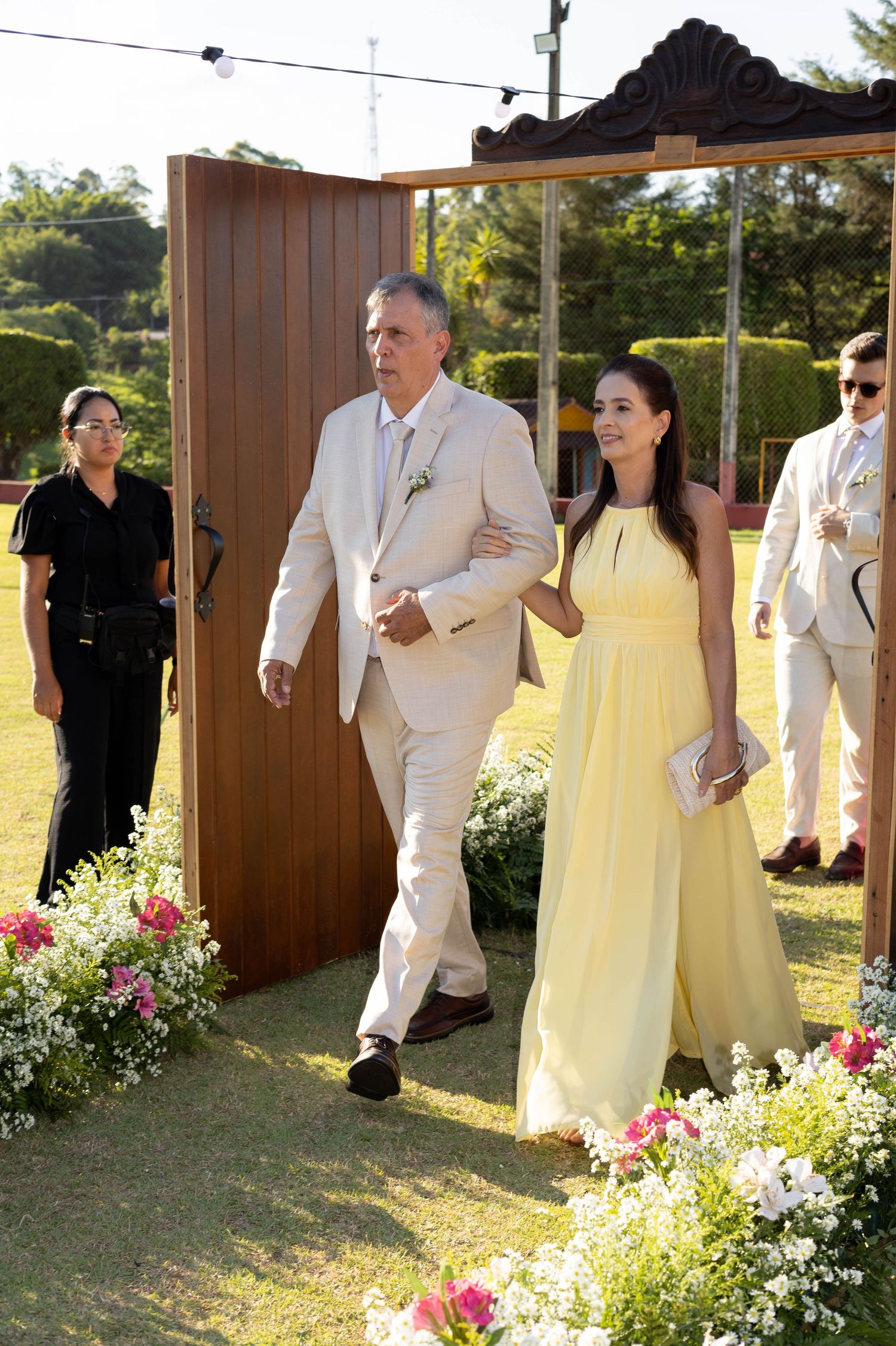 Elegant couple in yellow gown and beige suit walking through floral archway at outdoor wedding ceremony.