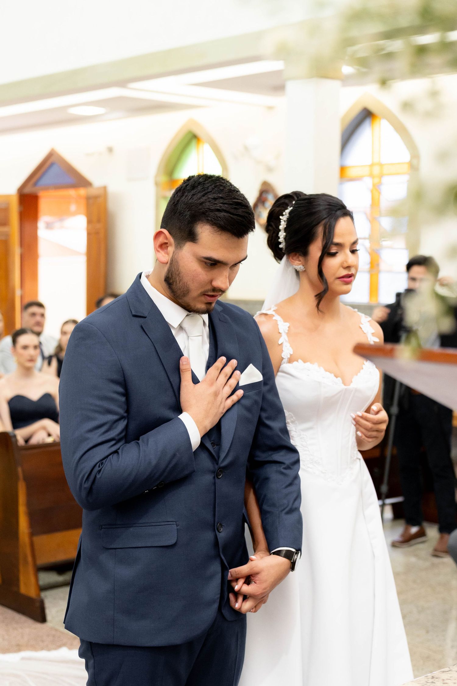 Bride and groom praying together at church altar during wedding ceremony, groom in navy suit, bride in white lace gown.