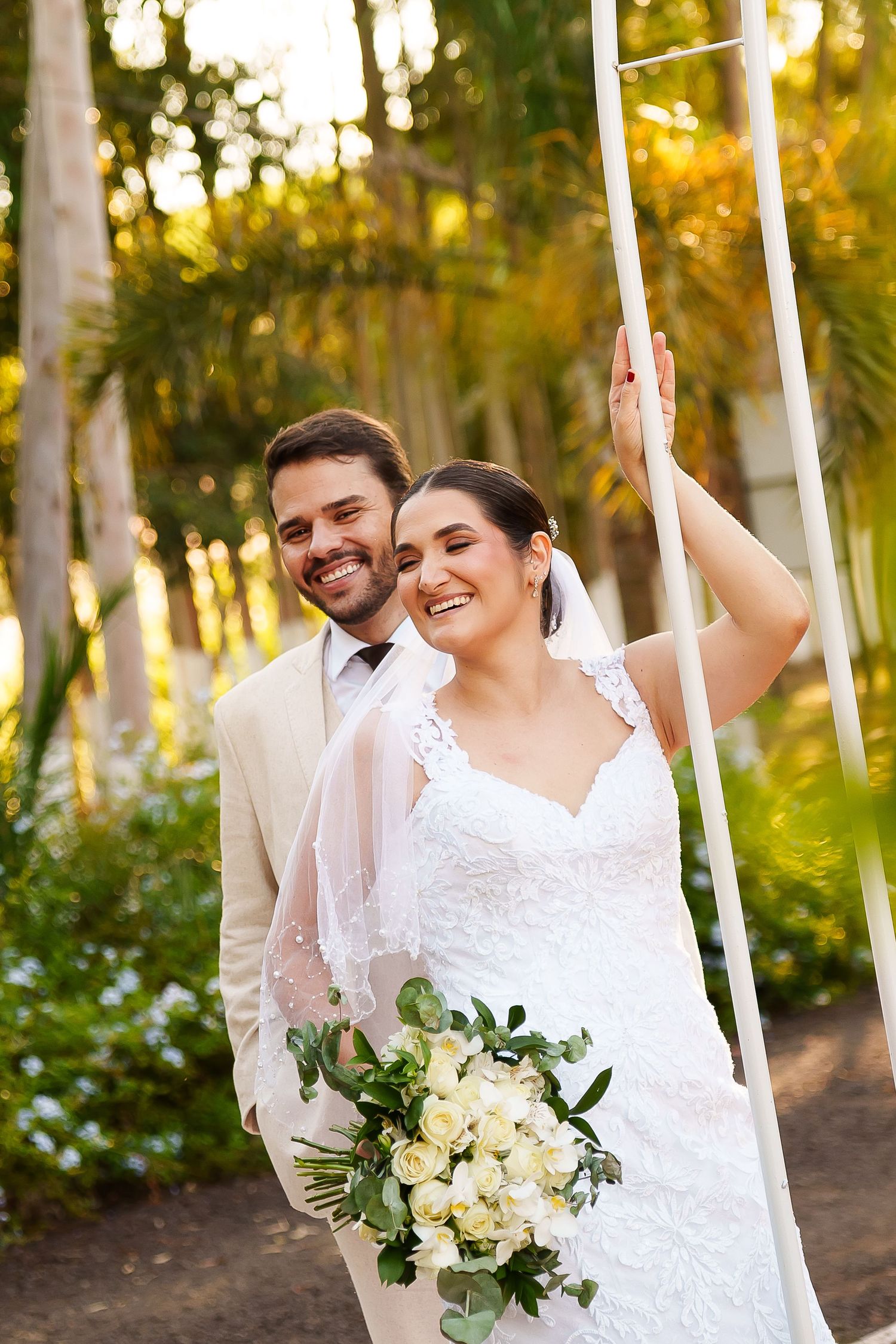 Smiling bride in white lace gown holding white roses bouquet with groom in beige suit in lush garden setting.