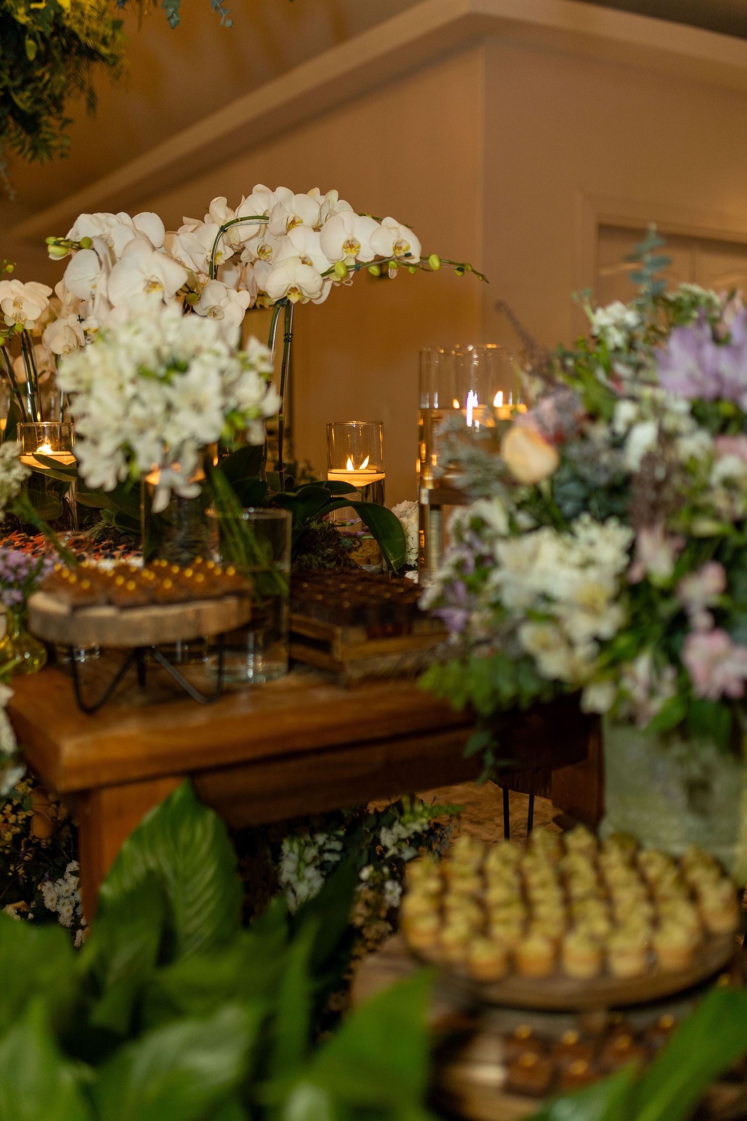 Elegant wedding dessert table with white orchids, candles, and golden treats amid lush greenery.