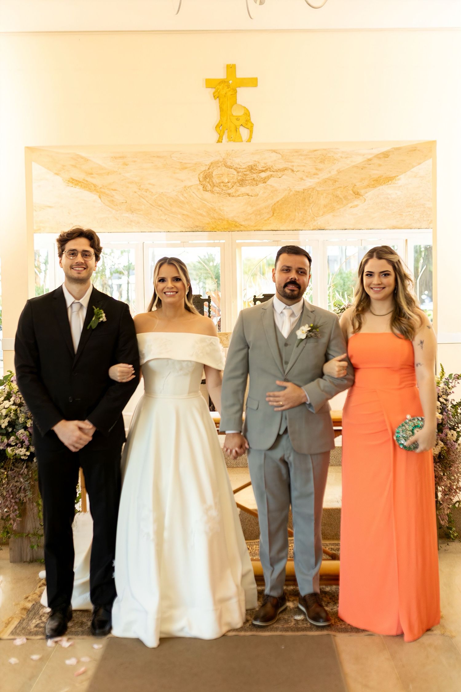 Wedding party posing in church, bride in white gown, groom in gray suit, groomsman in black tux, bridesmaid in orange dress.