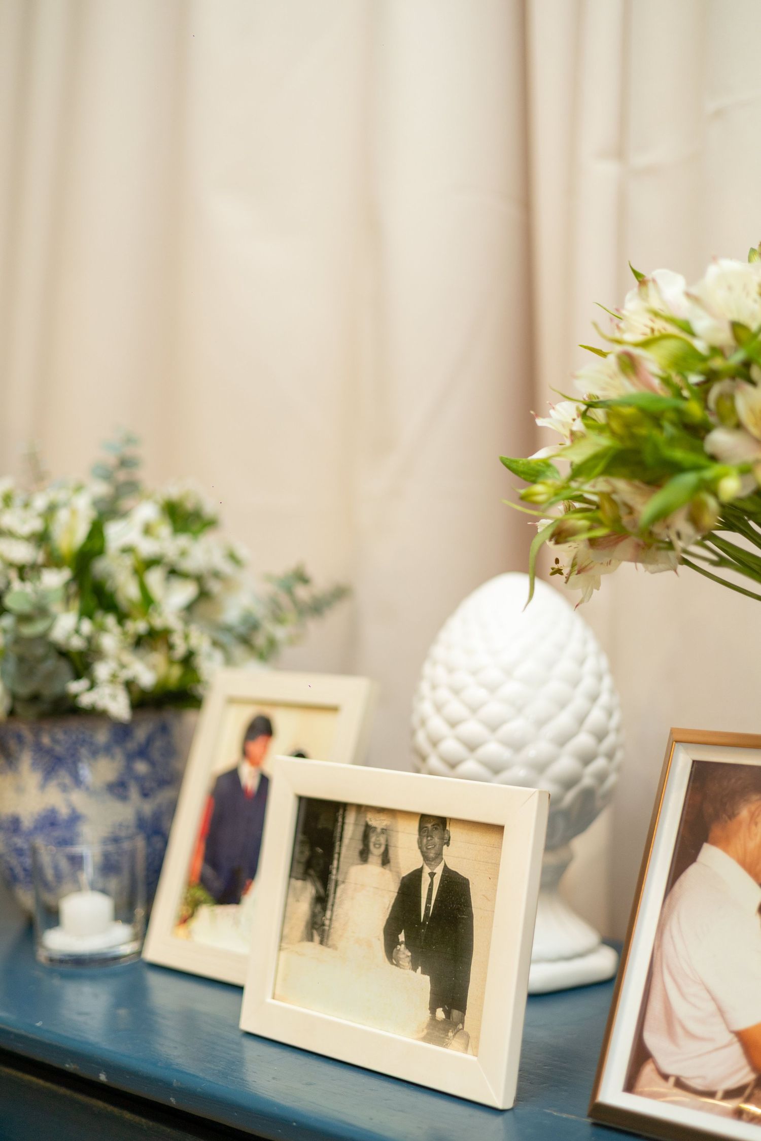Framed family photos on a blue table with white floral arrangements and a ceramic vase in a warm interior setting.