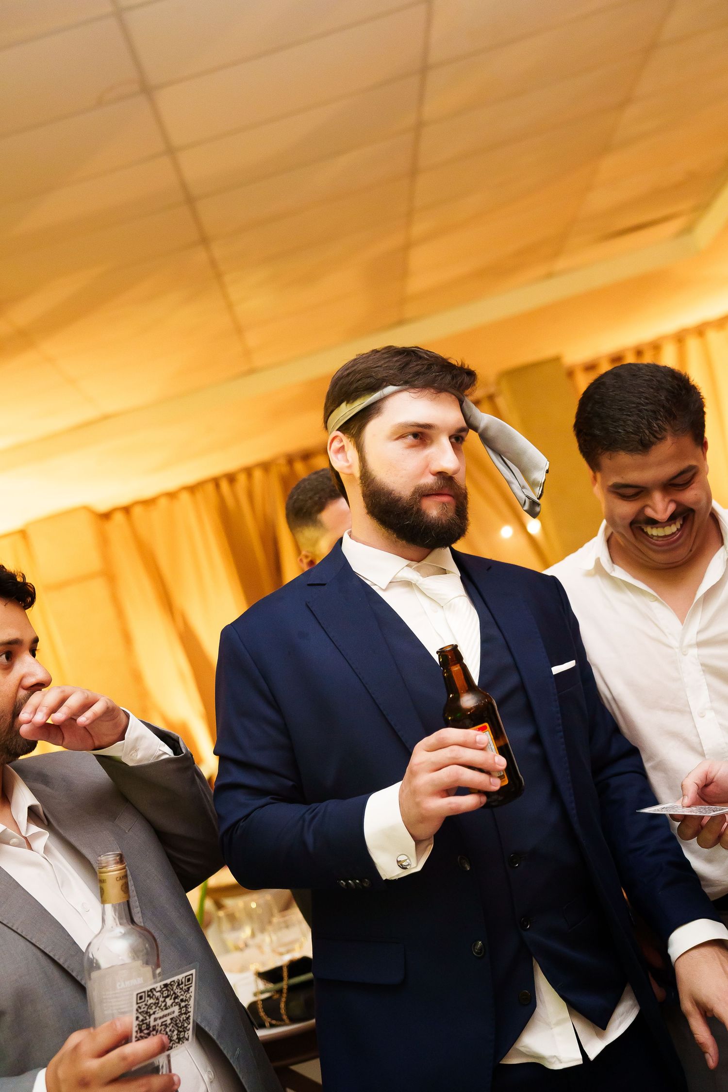 Bearded groom in navy suit holds beer bottle at wedding reception while friends laugh nearby.