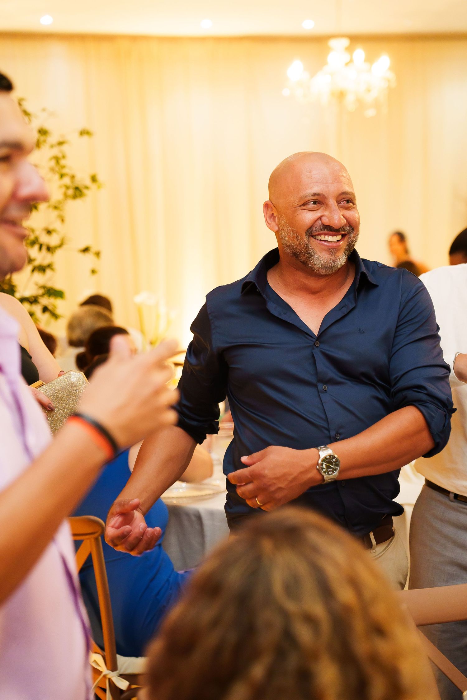 Bald man with beard smiling joyfully at an elegant indoor celebration, wearing a navy blue shirt and watch.