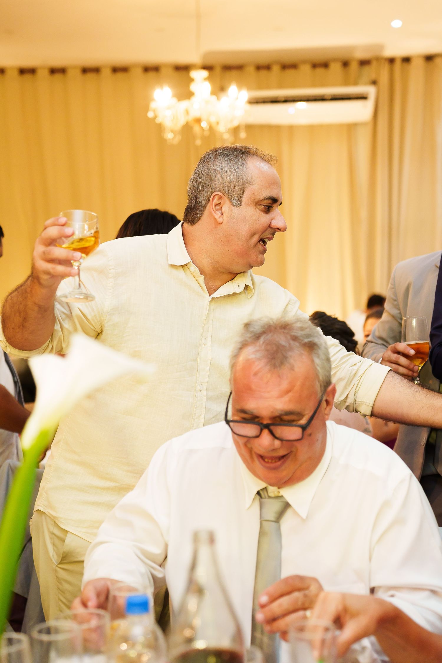 Two men celebrating at a formal event, one standing holding a whiskey glass, the other seated wearing glasses and a tie.