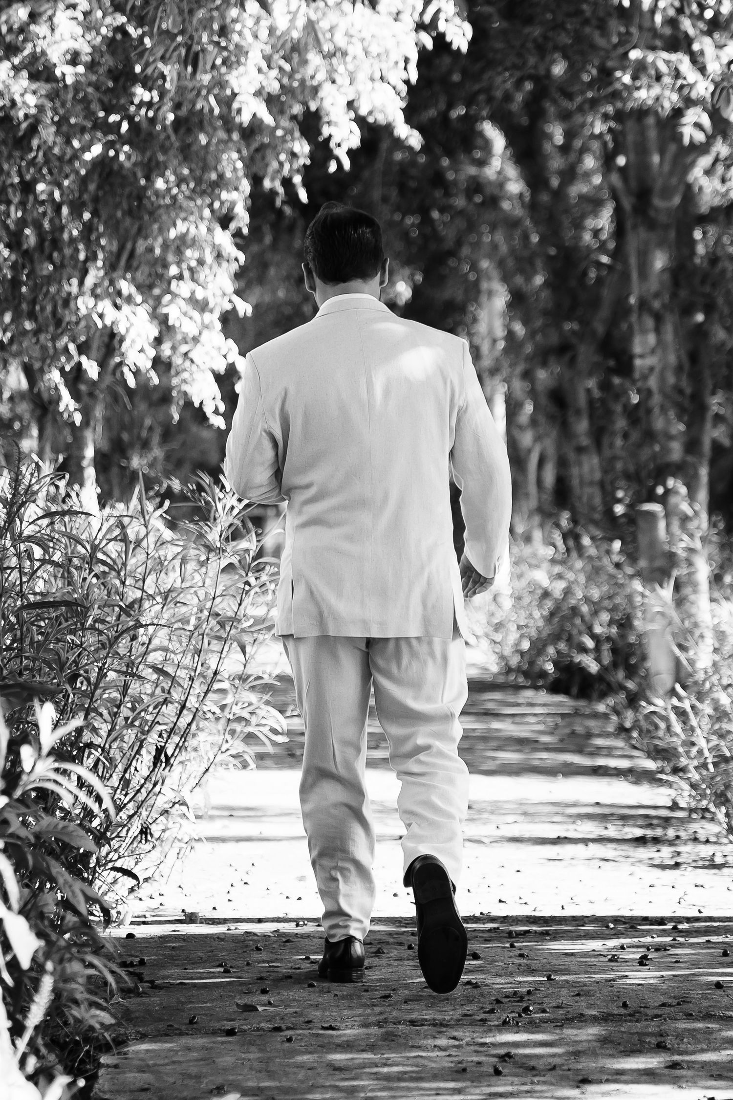 Black and white photo of a man in white suit walking away down a tree-lined path.