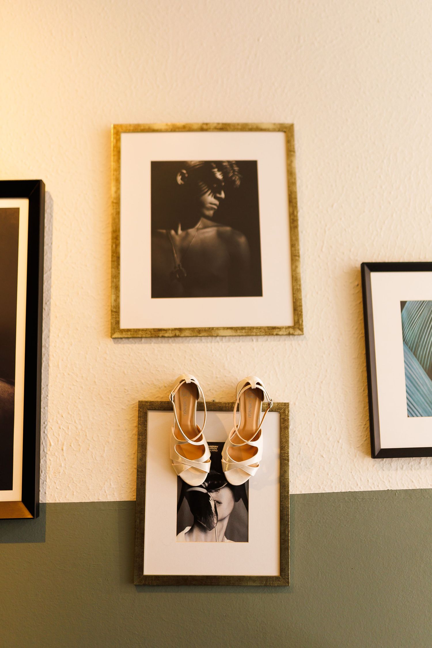 White strappy heels hanging on a gold picture frame beside black and white portrait photographs on a green and white wall.