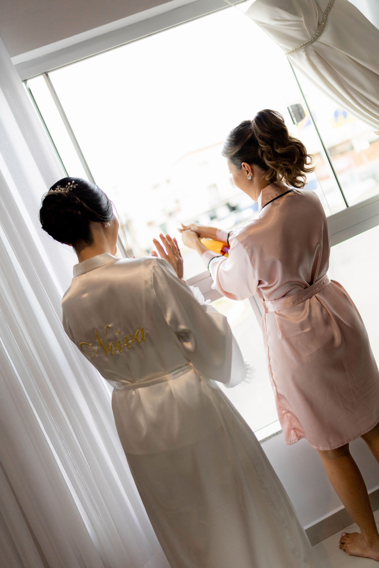 Bride and bridesmaid in silk robes preparing together by a bright window on wedding morning.