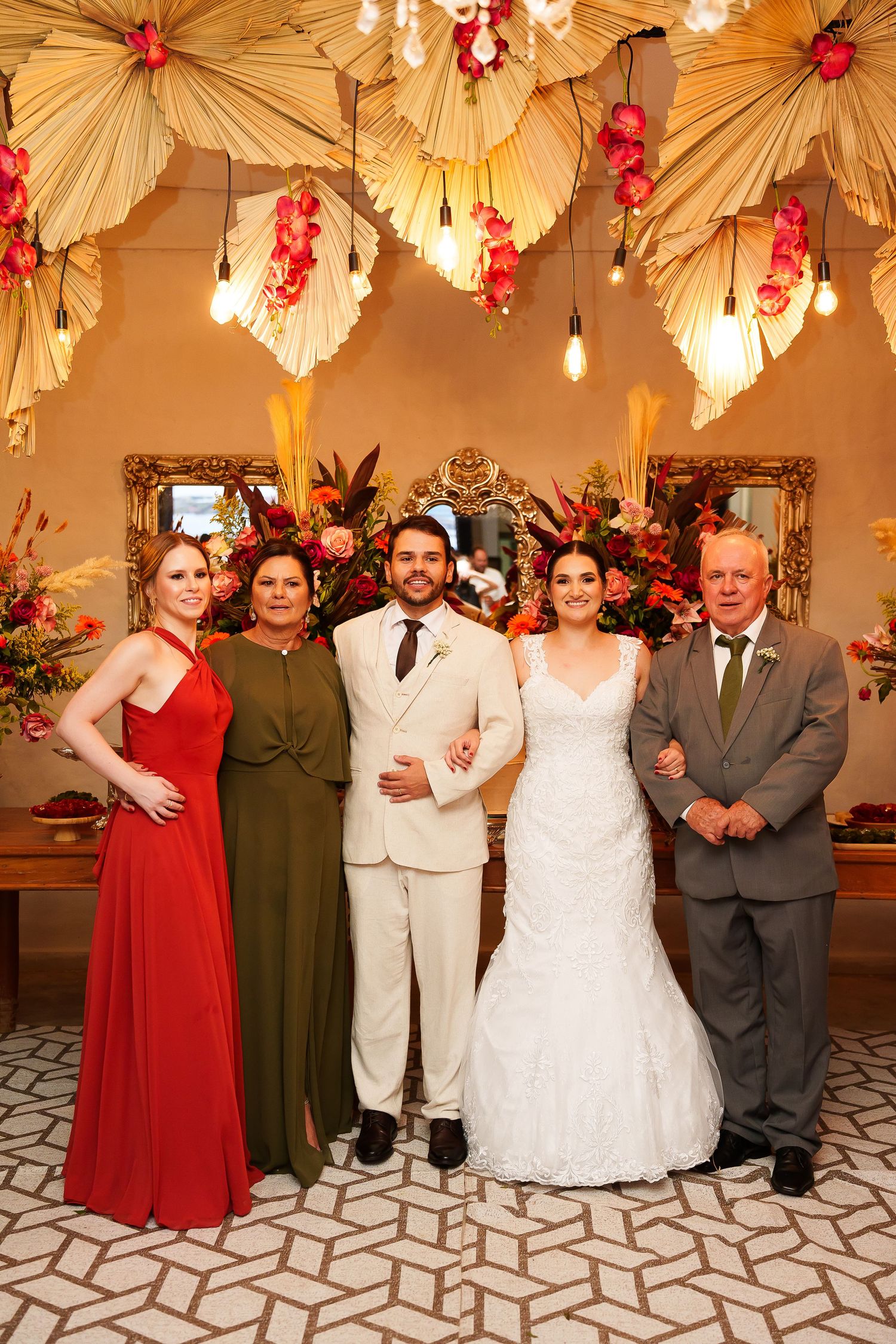 Wedding party of five posing indoors with floral decor, bride in white gown, groom in beige suit, guests in formal attire.