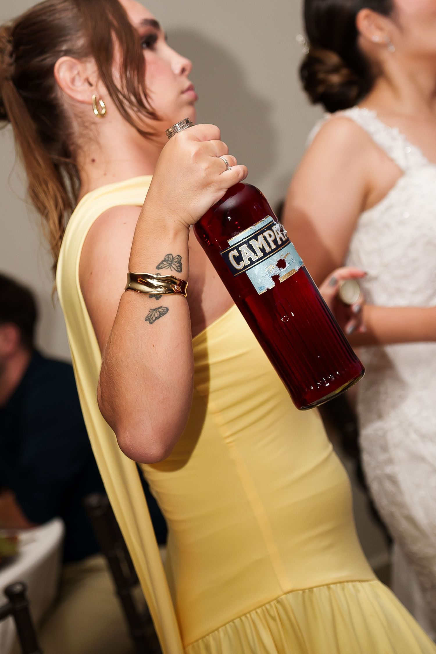 Woman in yellow dress holding a Campari bottle at a social event, showing tattoos and gold bracelets.