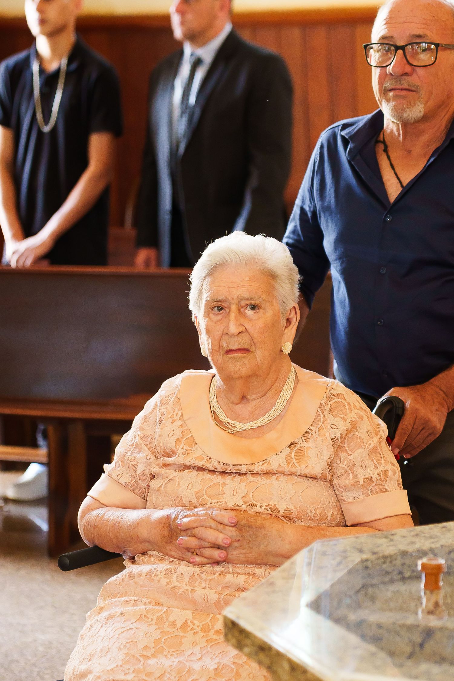 Elderly woman in lace dress seated at marble table, with two men standing behind her in formal indoor setting.