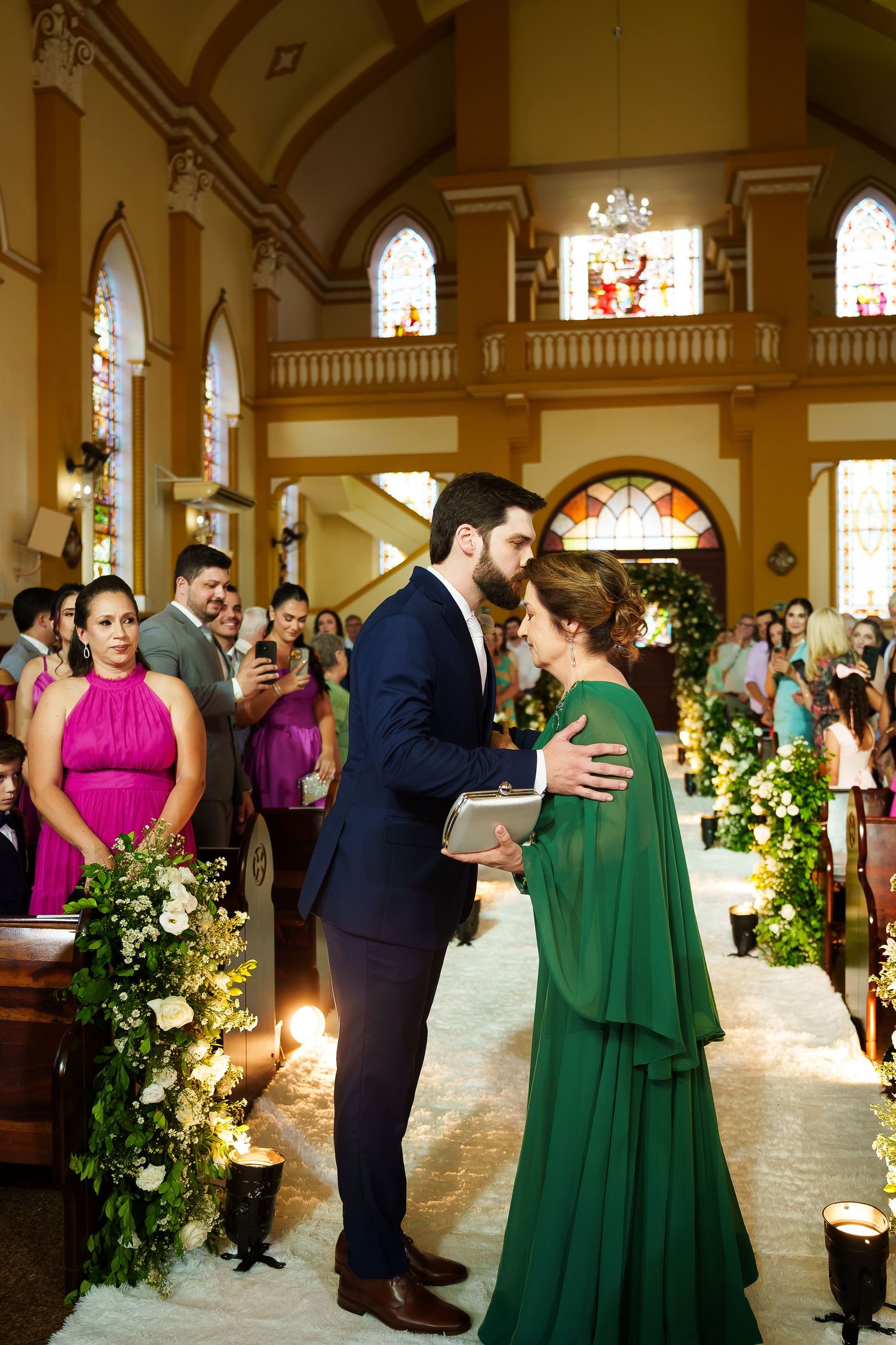 Groom in navy suit greets woman in green cape dress at church wedding ceremony aisle with floral arrangements.