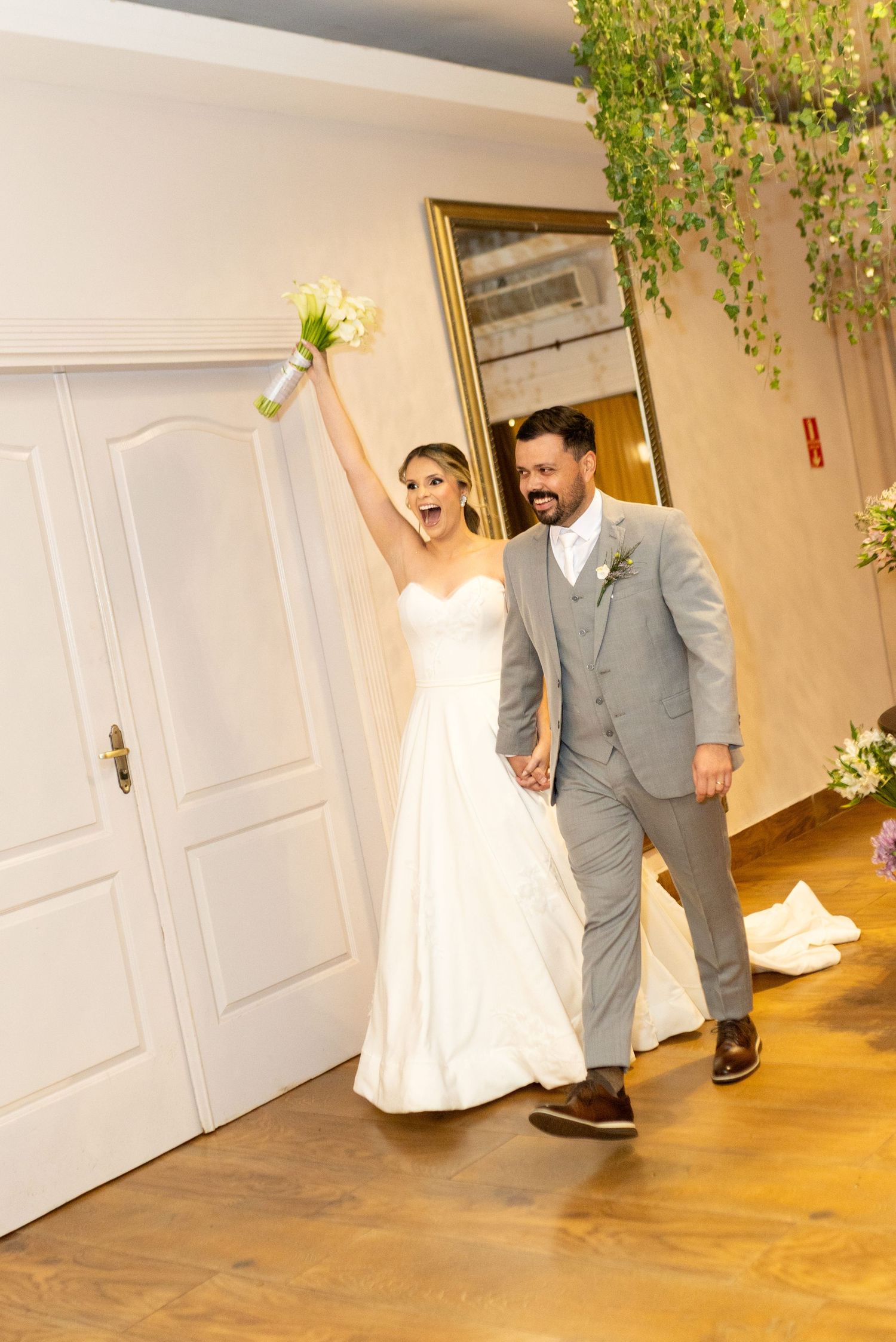 Joyful bride in white gown raises bouquet while groom in gray suit smiles during wedding reception entrance.