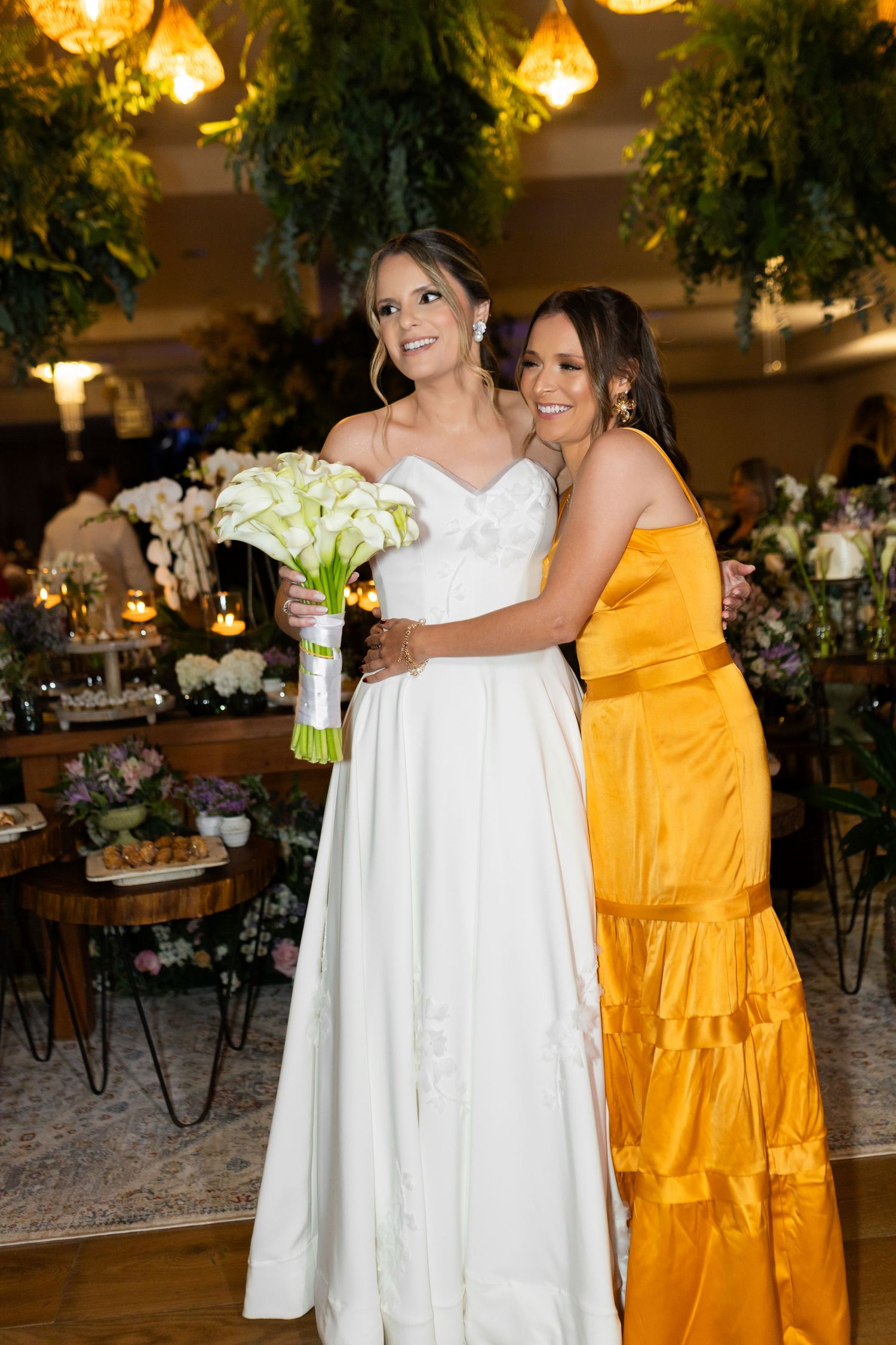 Bride in white gown holding calla lily bouquet poses with woman in golden yellow dress at elegant wedding reception.
