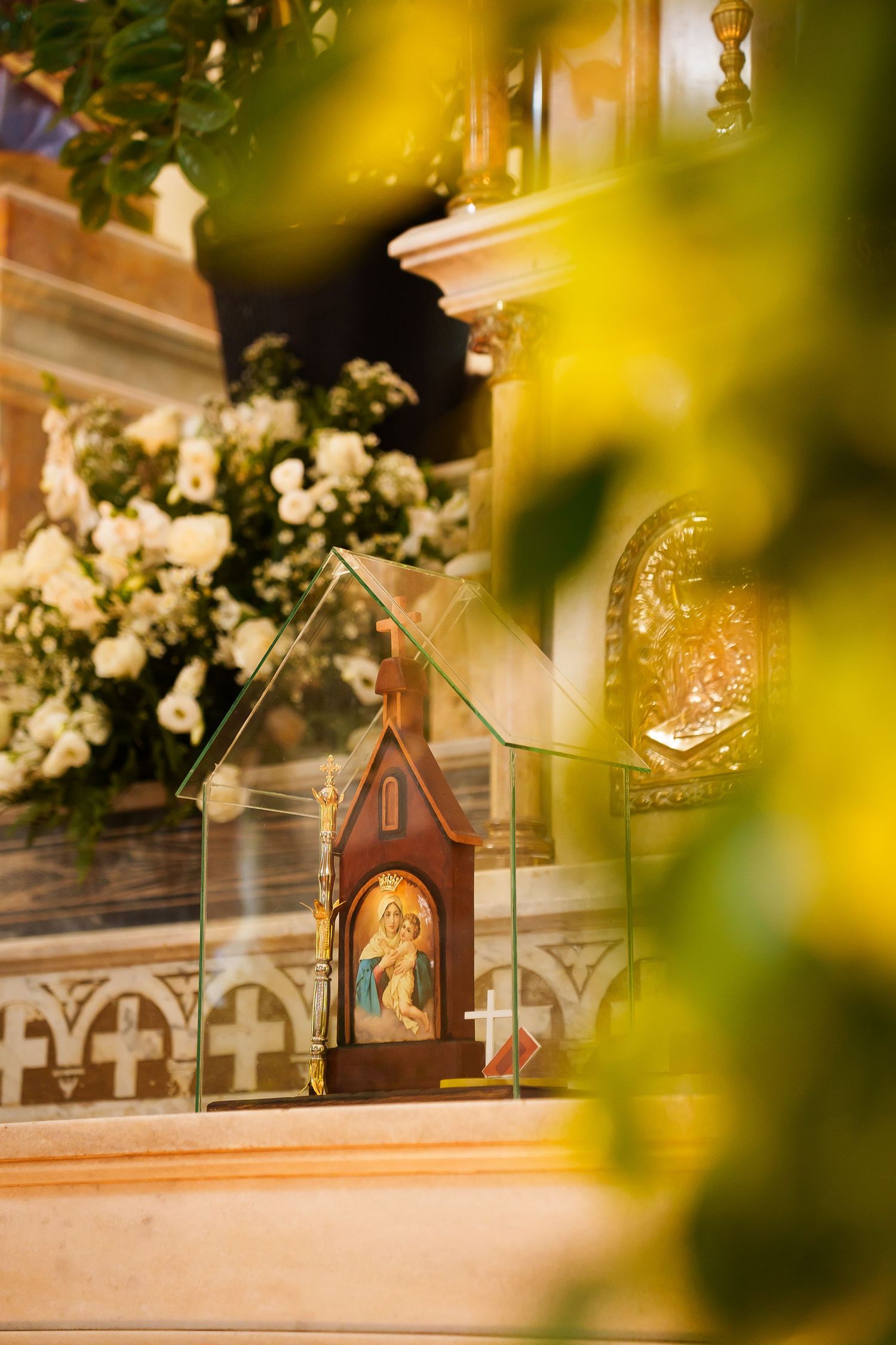 Orthodox Christian icon in decorative wooden shrine with glass panels, surrounded by white flowers in ornate church interior.