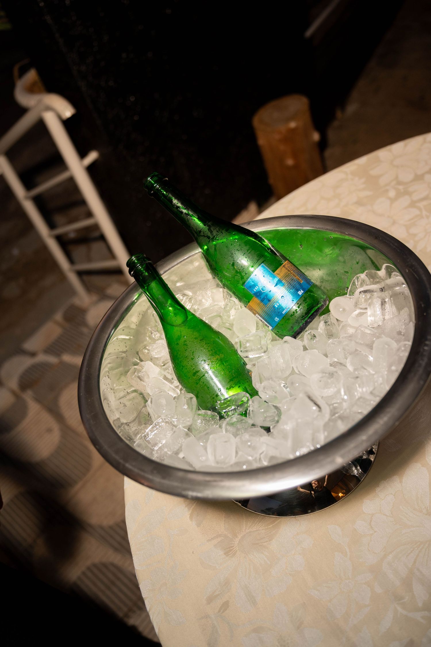 Two green bottles chilling in a silver ice bucket filled with ice cubes on a white tablecloth table.