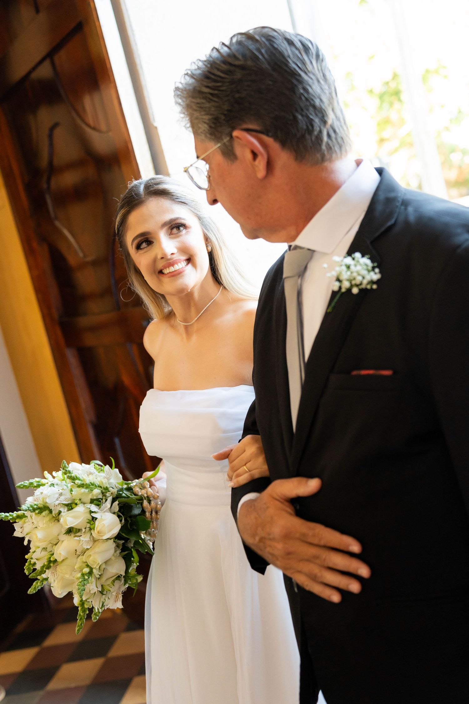 Bride in white strapless gown holding bouquet smiles at older man in dark suit with boutonniere at wedding ceremony.