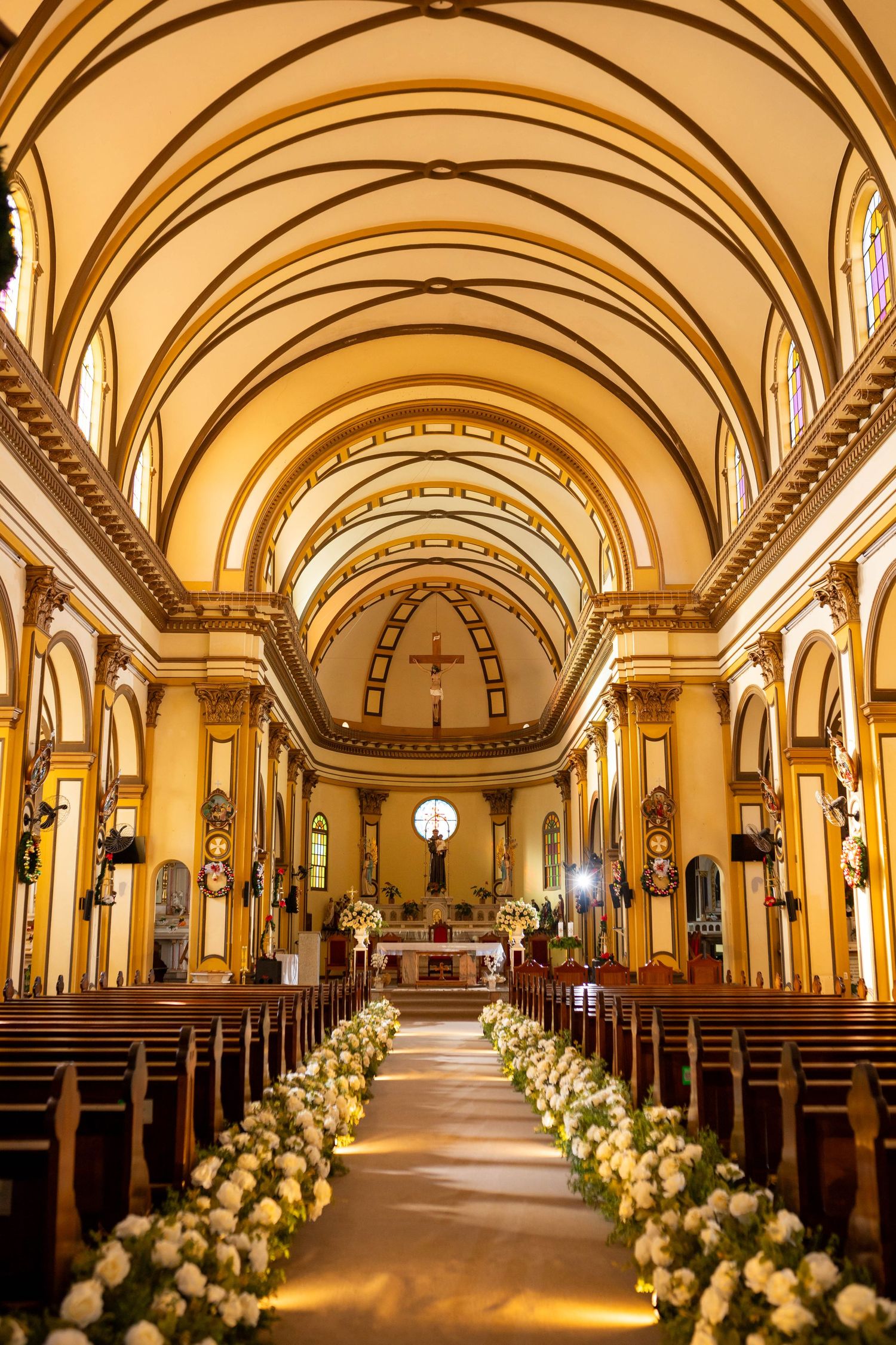 Elegant church interior with vaulted golden ceilings, wooden pews, and white floral aisle decorations leading to an ornate altar.