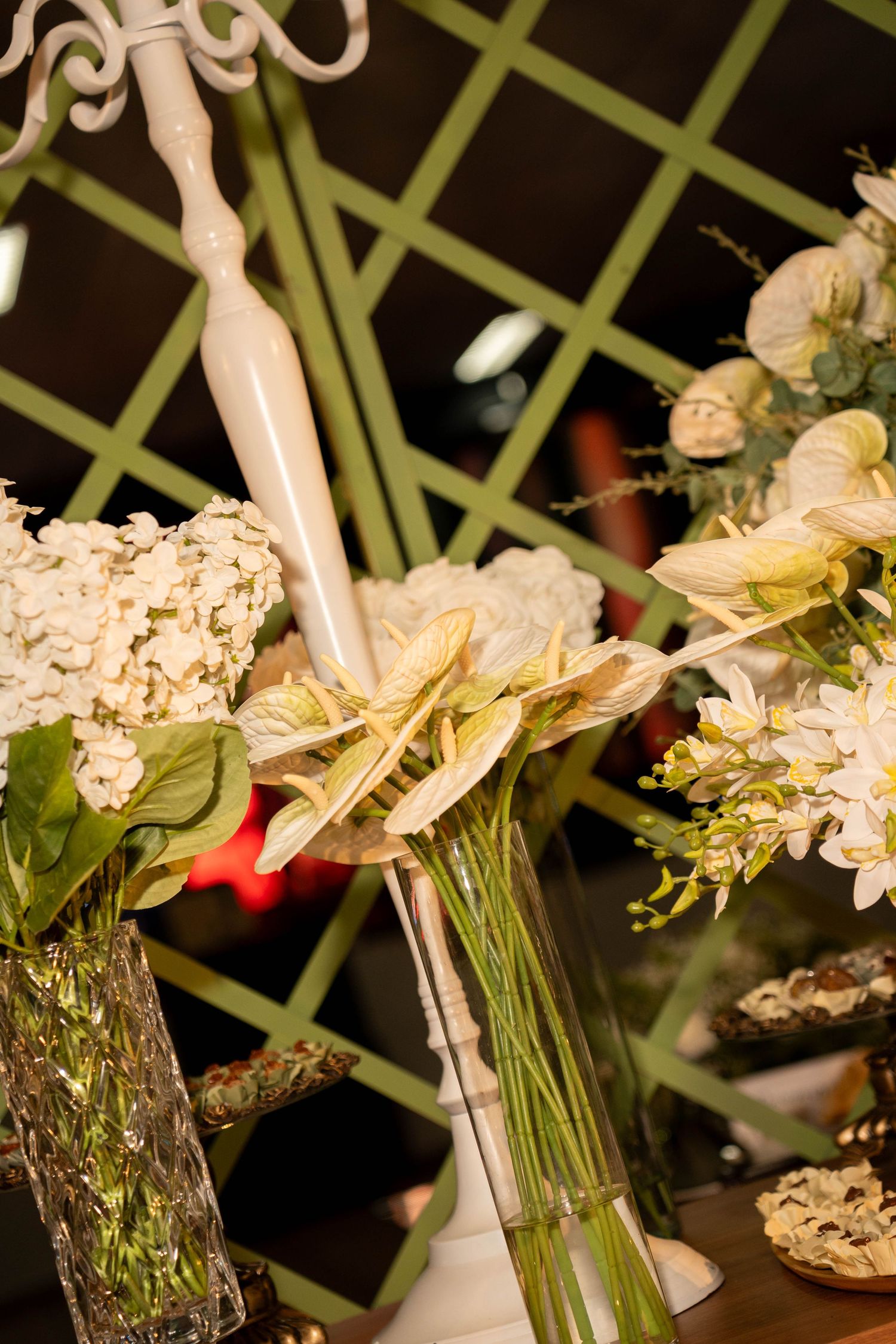 Elegant floral arrangement with white hydrangeas and anthurium in glass vases on a table with green lattice backdrop.