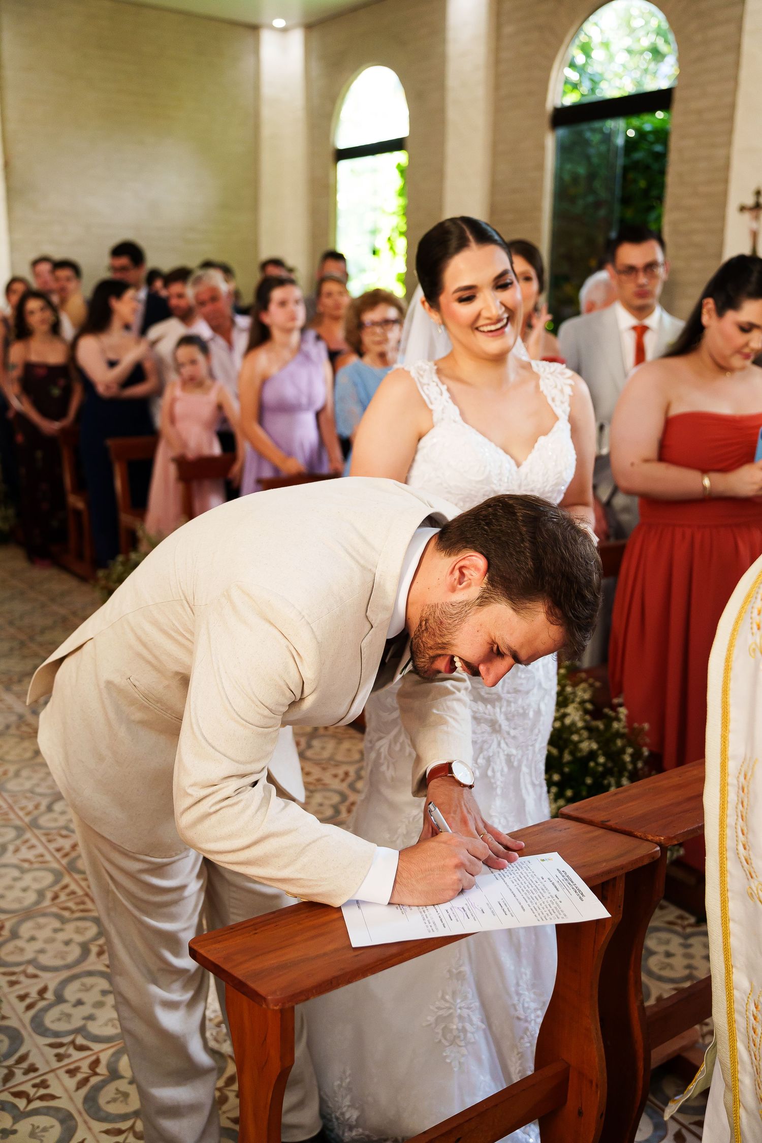 Groom signing wedding certificate at church altar while smiling bride in white lace dress watches guests celebrate.