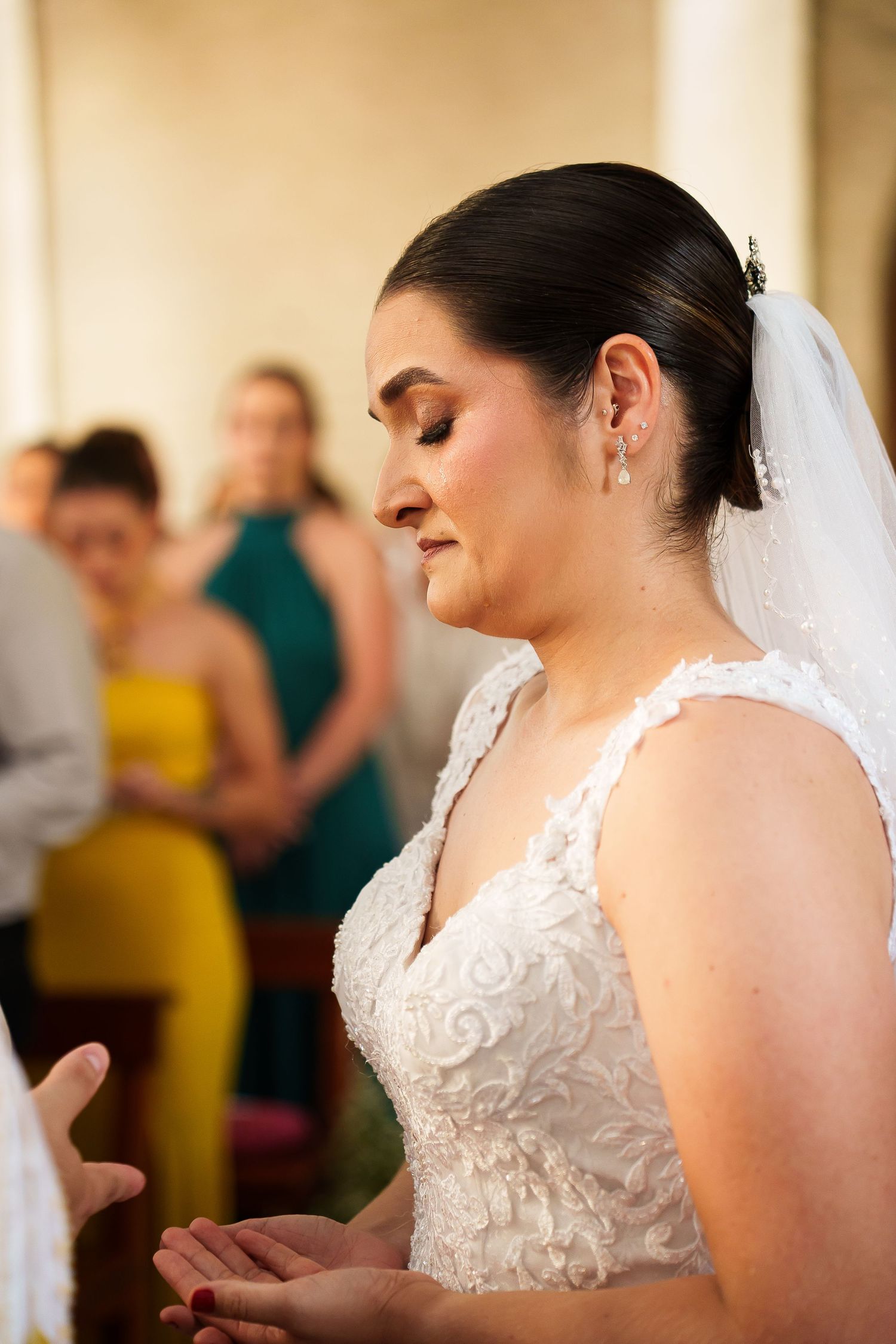Bride in white lace gown and veil with eyes closed during wedding ceremony, guests in colorful attire in background.