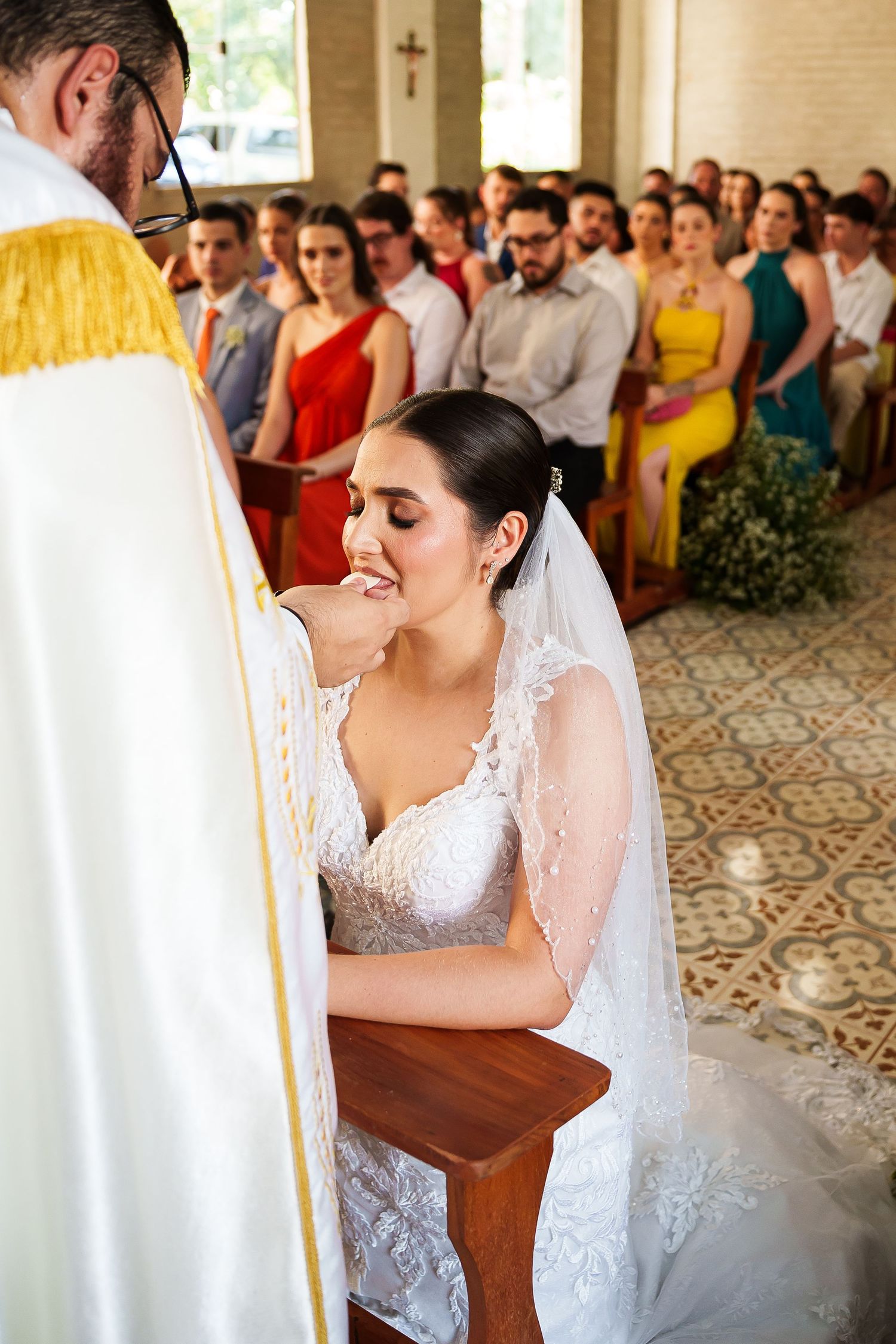 Bride in white lace gown and veil kneels at altar during Catholic wedding ceremony with guests seated in pews behind her.