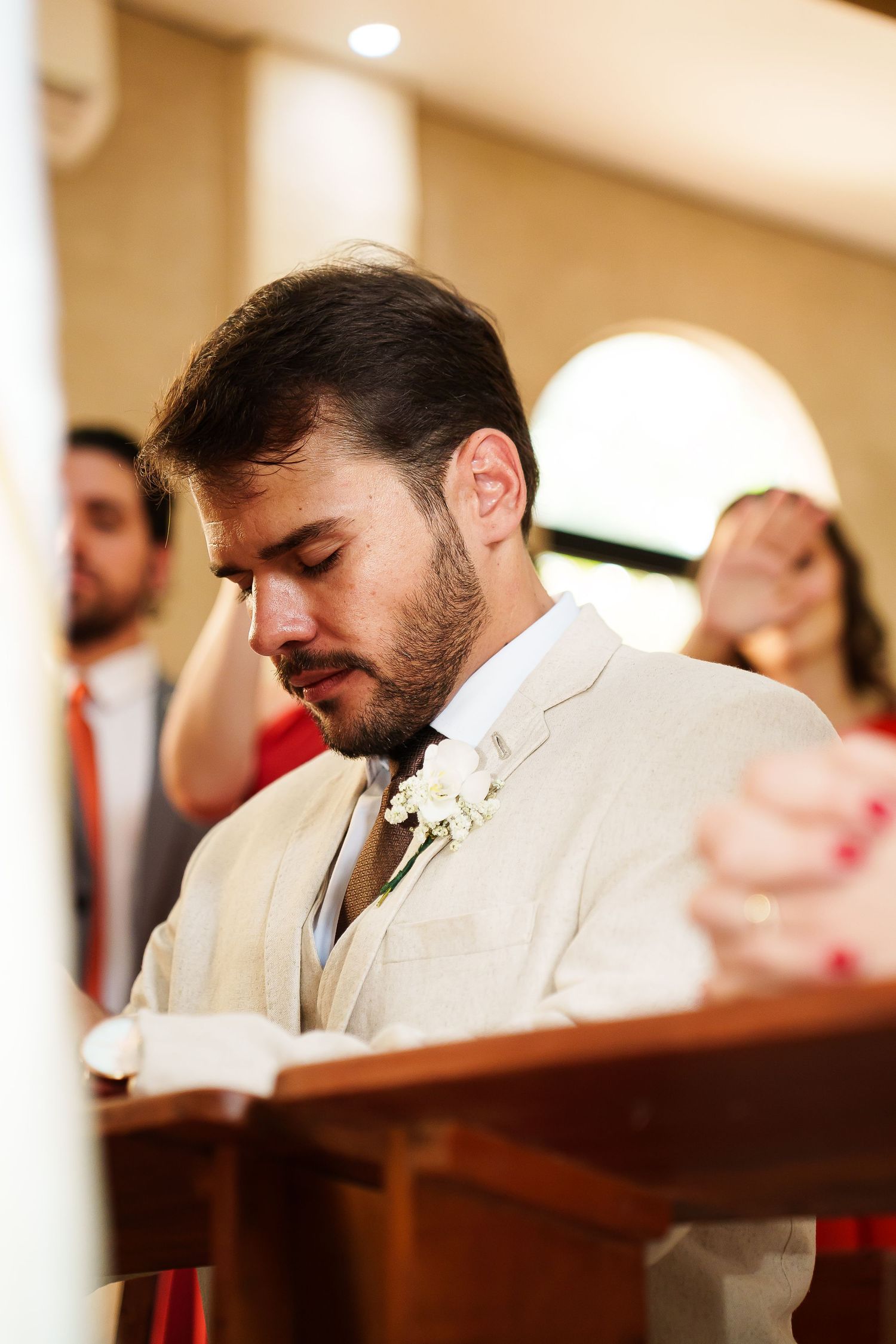 Groom in beige suit with white boutonniere bows head in prayer at church altar during wedding ceremony.