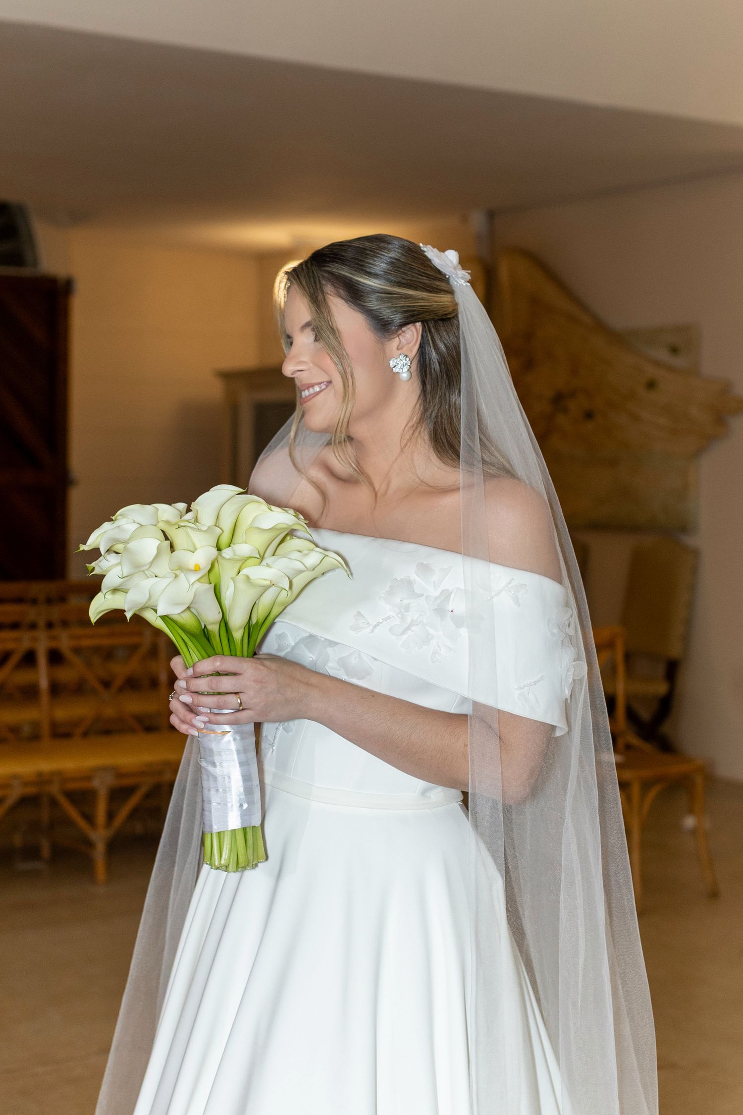 Smiling bride in off-shoulder white gown holds calla lily bouquet with veil draped elegantly indoors.