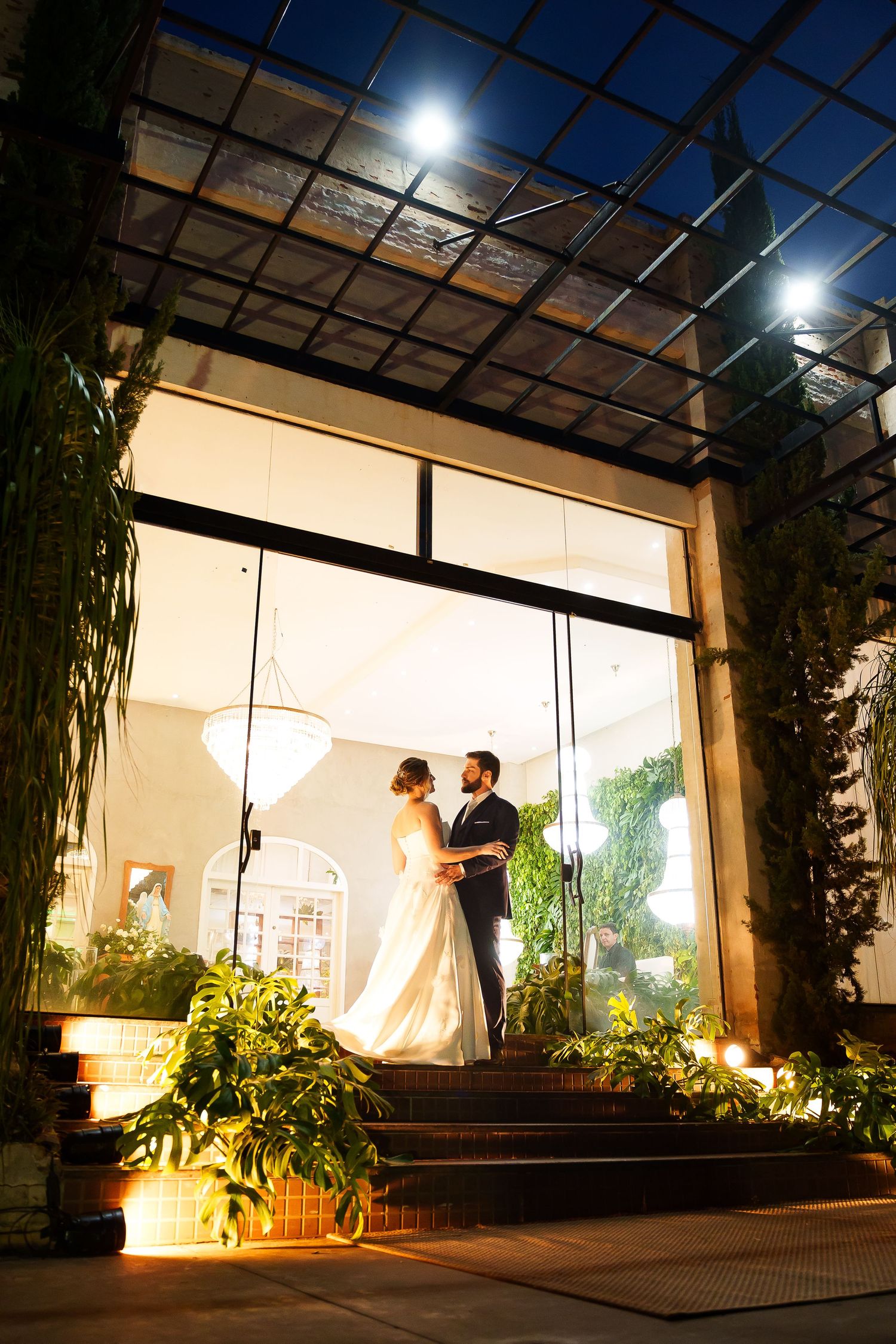 Bride and groom share a romantic embrace in a glowing glass doorway surrounded by lush greenery at night.