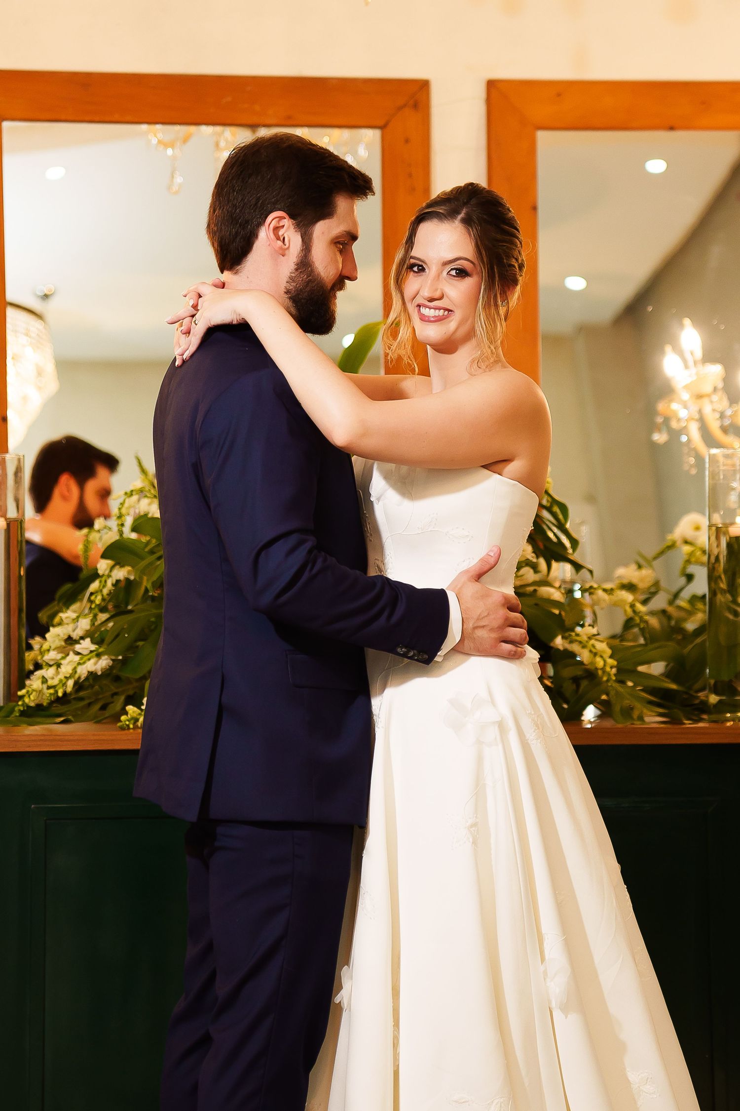 Bride and groom share first dance, bride in strapless white gown smiling, groom in navy suit, floral backdrop.