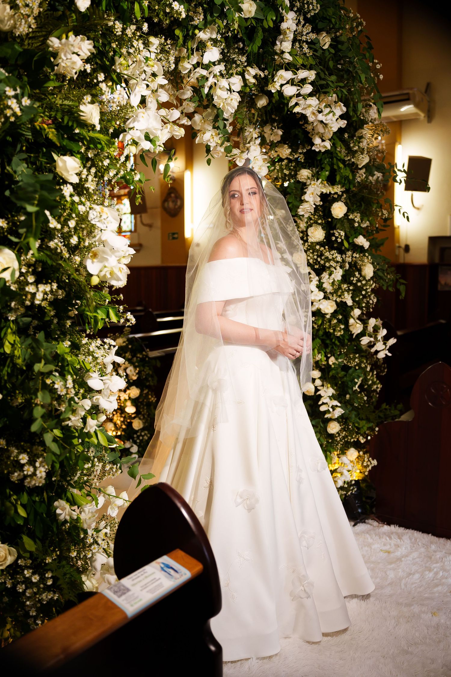 Bride in white off-shoulder gown and veil stands before lush floral arch with white roses in elegant church.