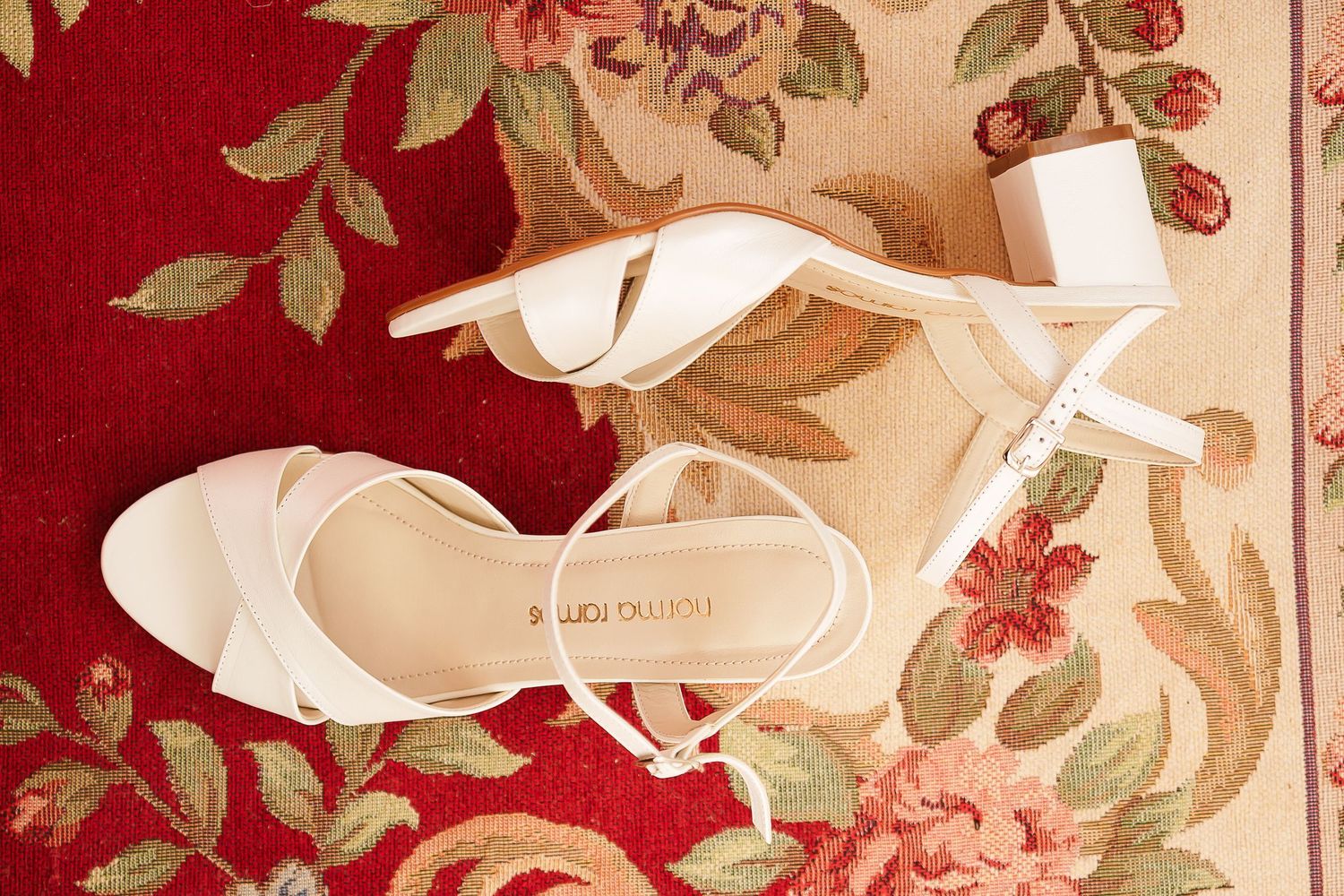 White strappy heeled sandals by Paula Olivia displayed on a floral red and cream area rug.