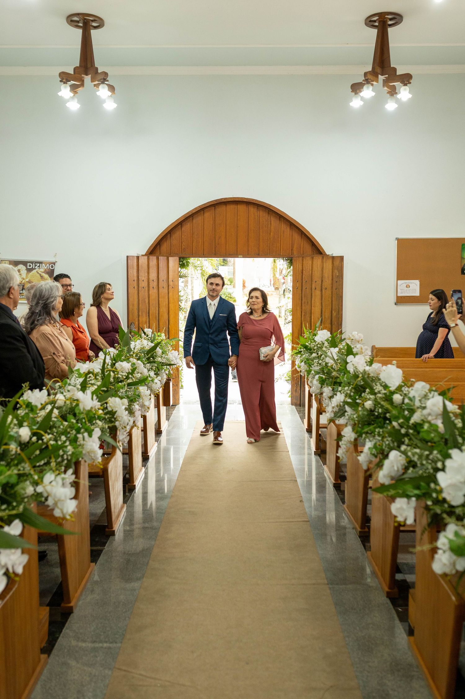 Groom and mother walk down a flower-lined church aisle during a wedding ceremony, with guests seated on wooden pews.
