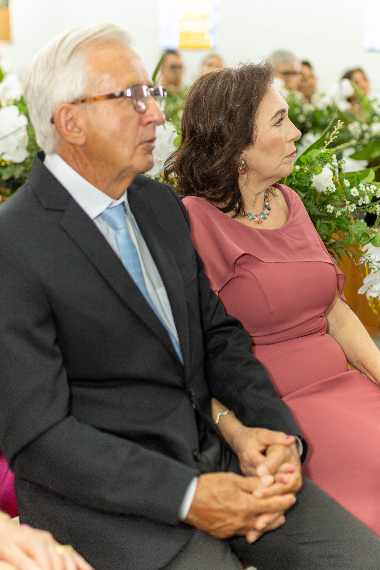 Elegant senior couple holding hands at a formal event, man in suit with tie, woman in pink dress with floral backdrop.