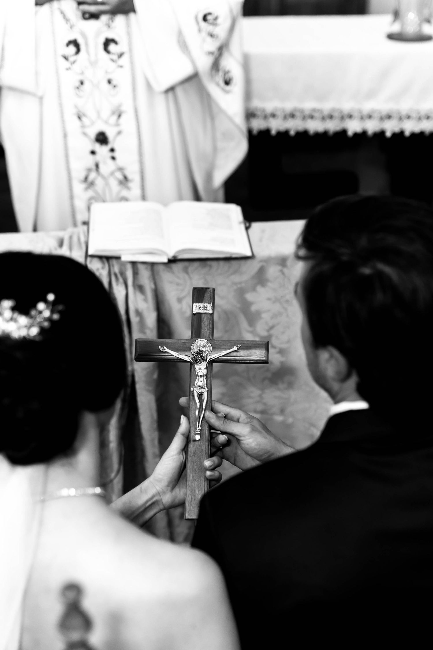 Black and white photo of a couple holding a crucifix during a Catholic wedding ceremony with a priest officiating at the altar.