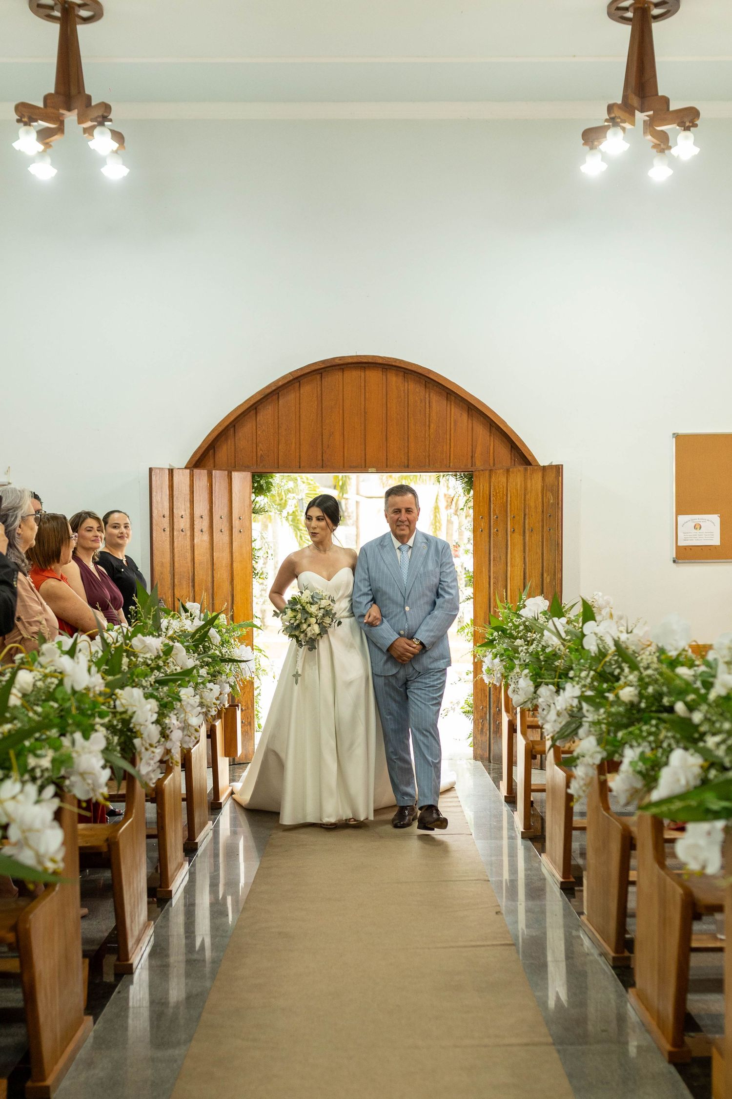 Bride walks down church aisle with escort, flanked by white floral arrangements on wooden pews during wedding ceremony.