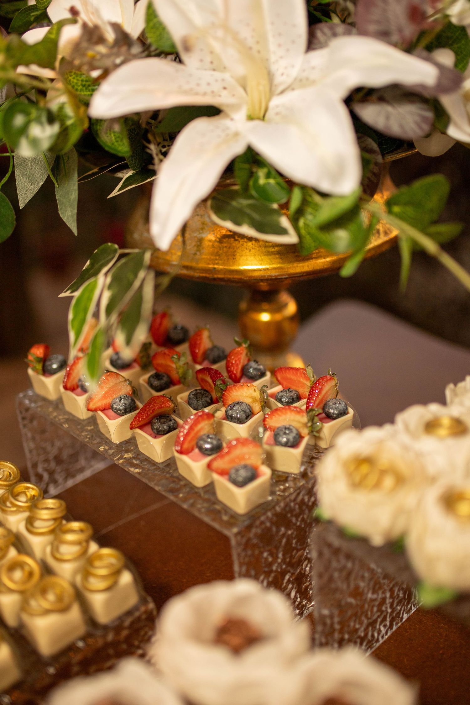 Elegant dessert table with fruit tarts topped with strawberries and blueberries, surrounded by white lily flowers at a wedding.