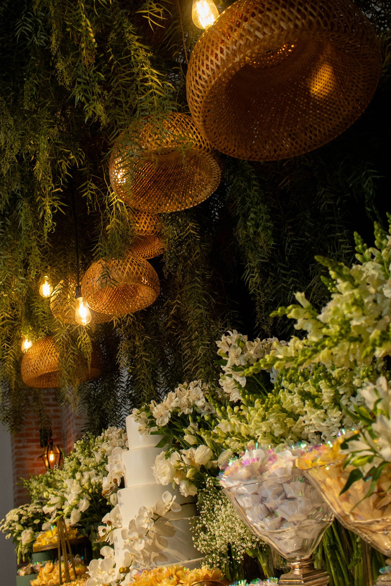 Elegant wedding dessert table with white flowers, tiered cake, wicker pendant lights, and lush greenery backdrop.