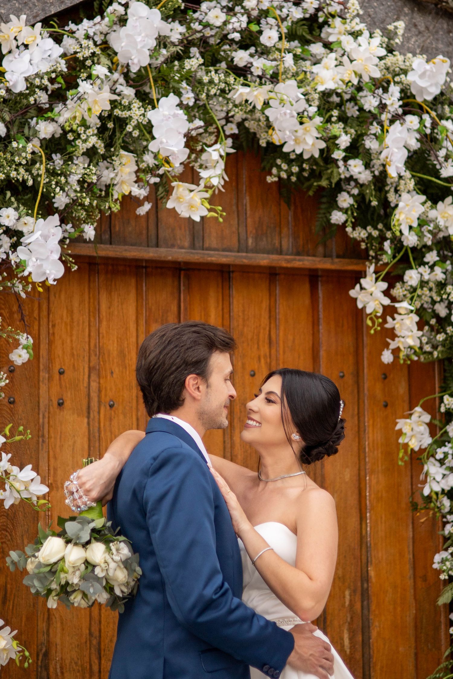 Bride and groom embrace under a white floral arch in front of a wooden door on their wedding day.