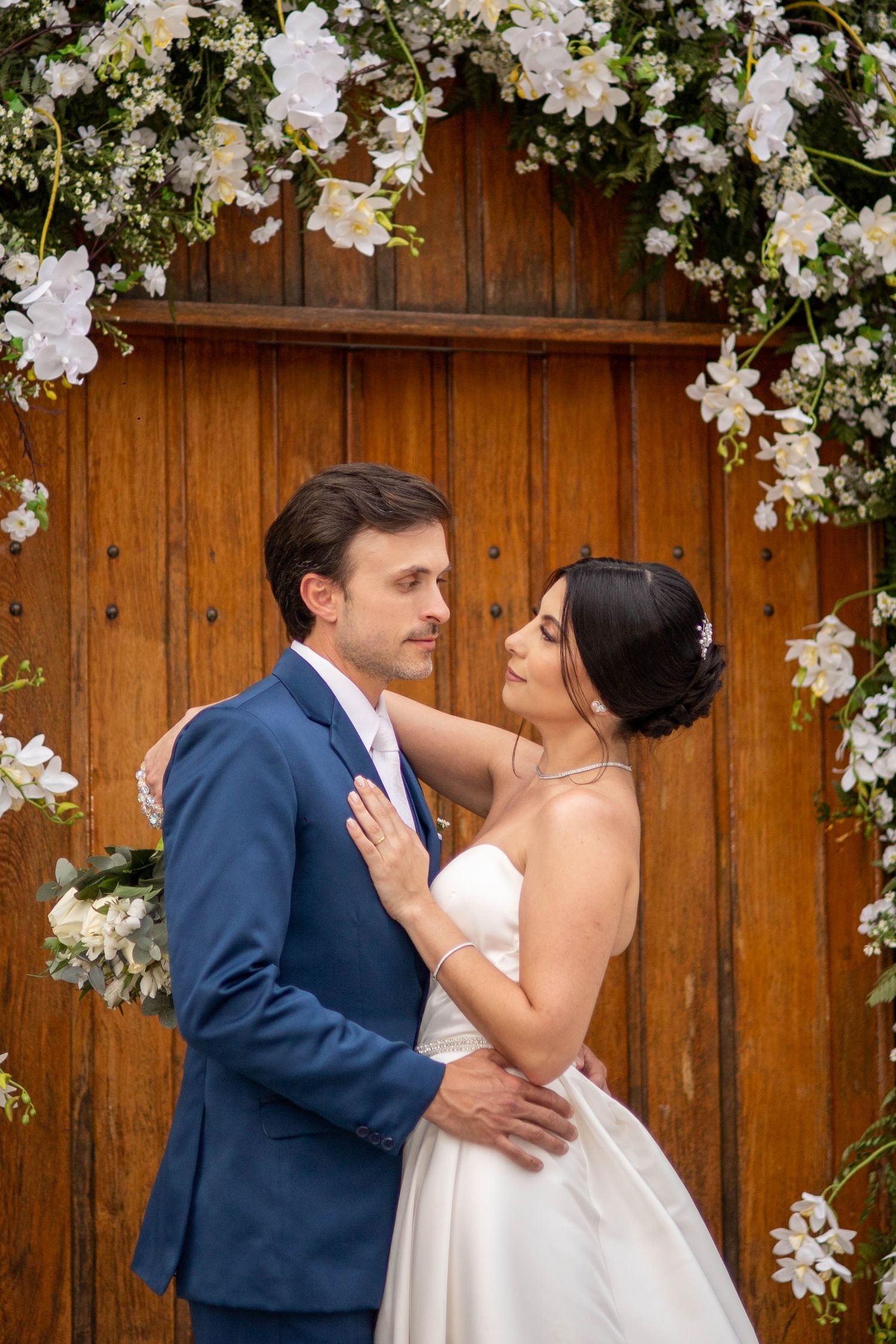 Bride and groom embrace under a floral arch with white flowers against a rustic wooden door backdrop.